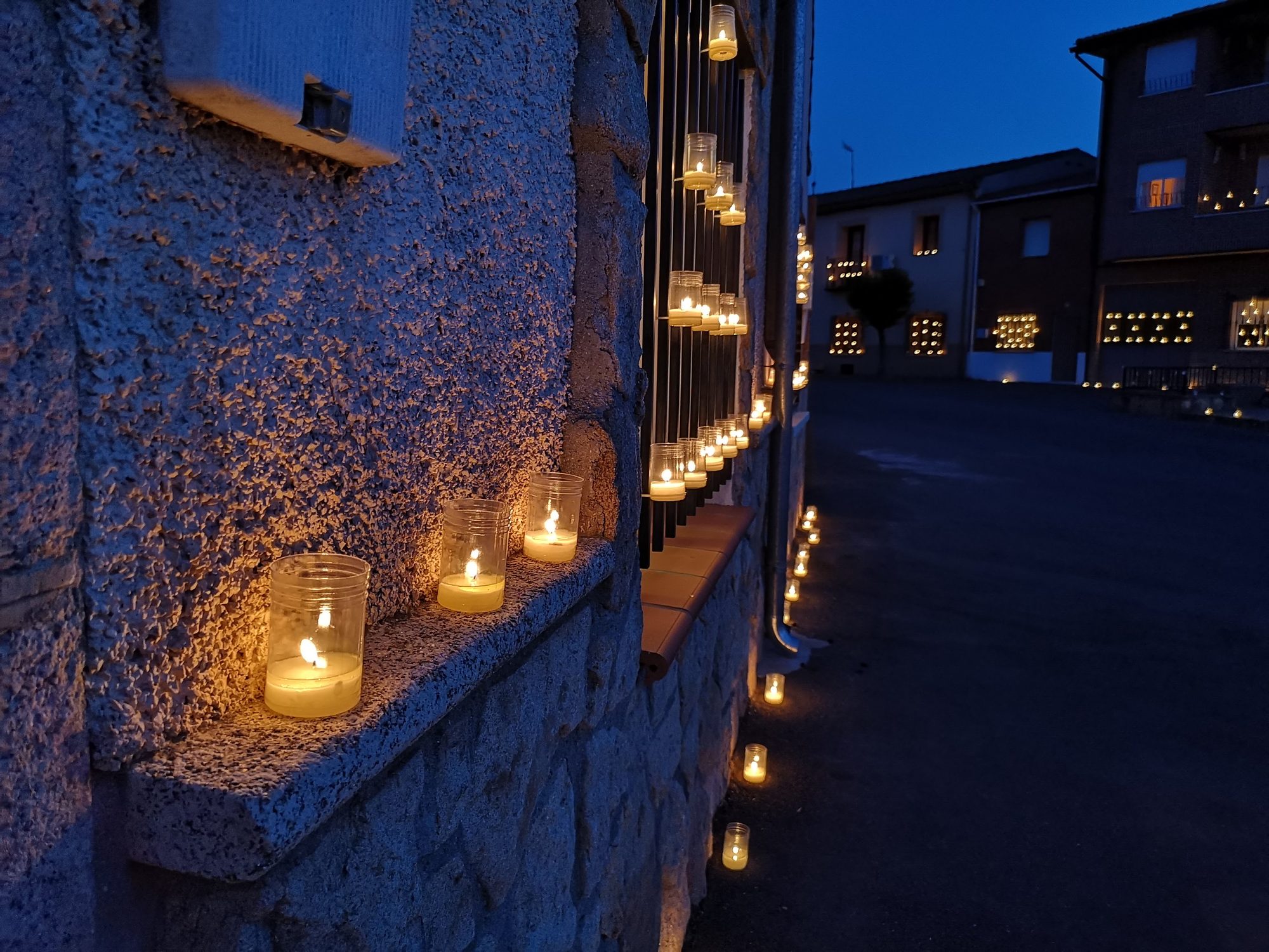 Velas en vasos de cristal iluminando ventanas y calles de un pueblo al anochecer.