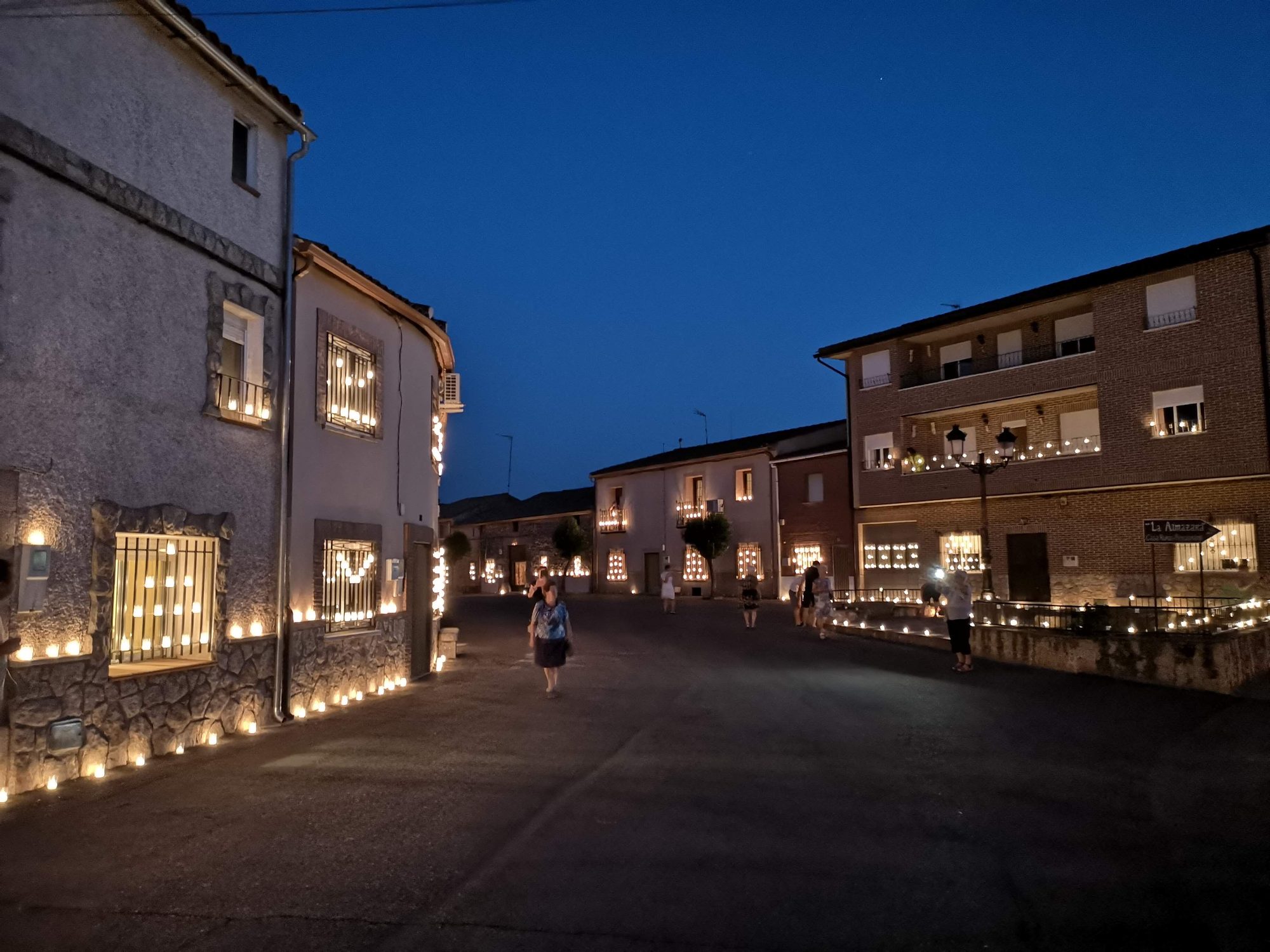 Calle de pueblo iluminada al anochecer por cientos de velas en ventanas y fachadas.