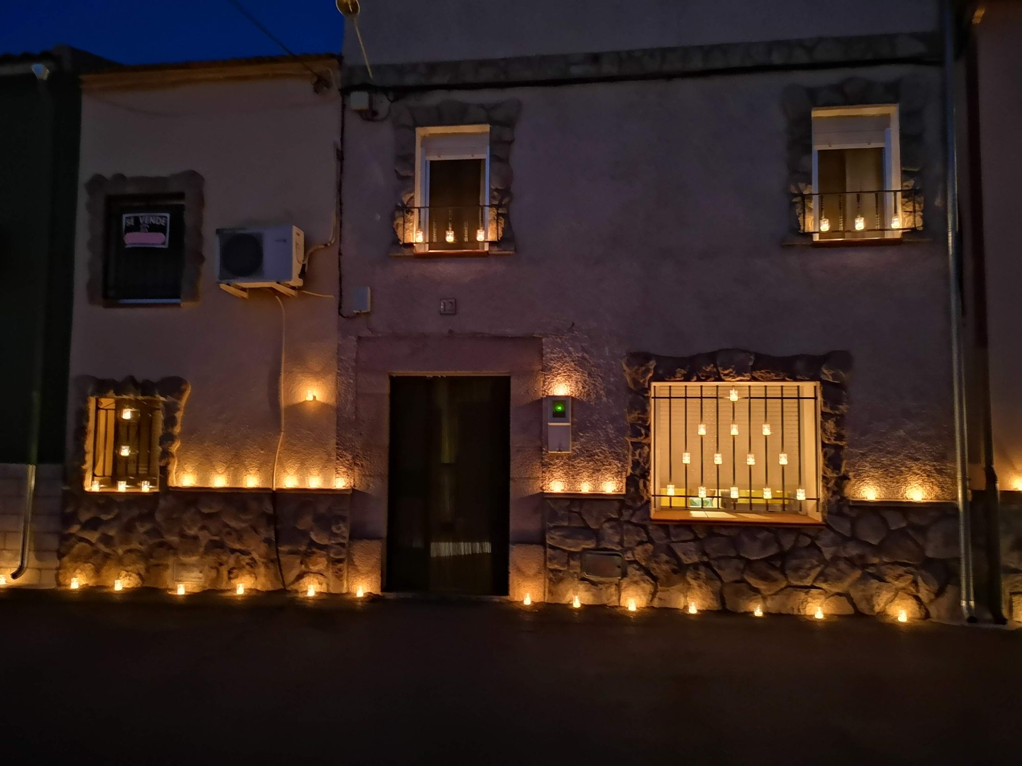 Fachada de una casa de pueblo iluminada de noche por numerosas velas encendidas.
