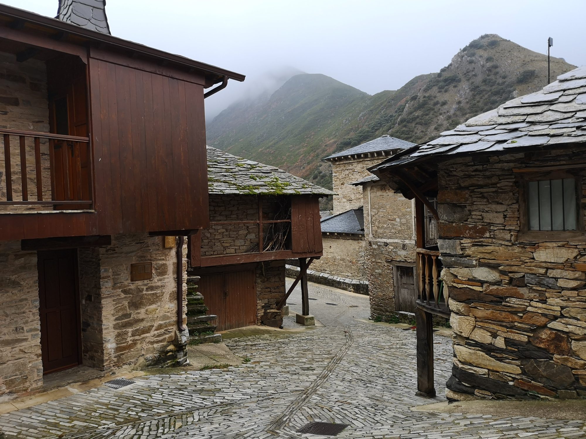 Calle empedrada con casas tradicionales de piedra y pizarra frente a montañas con niebla.