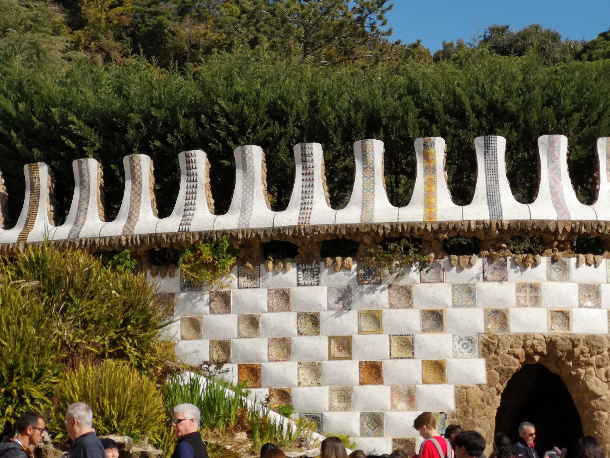Muro ondulado con mosaicos de cerámica trencadís en el Park Güell, rodeado de vegetación.