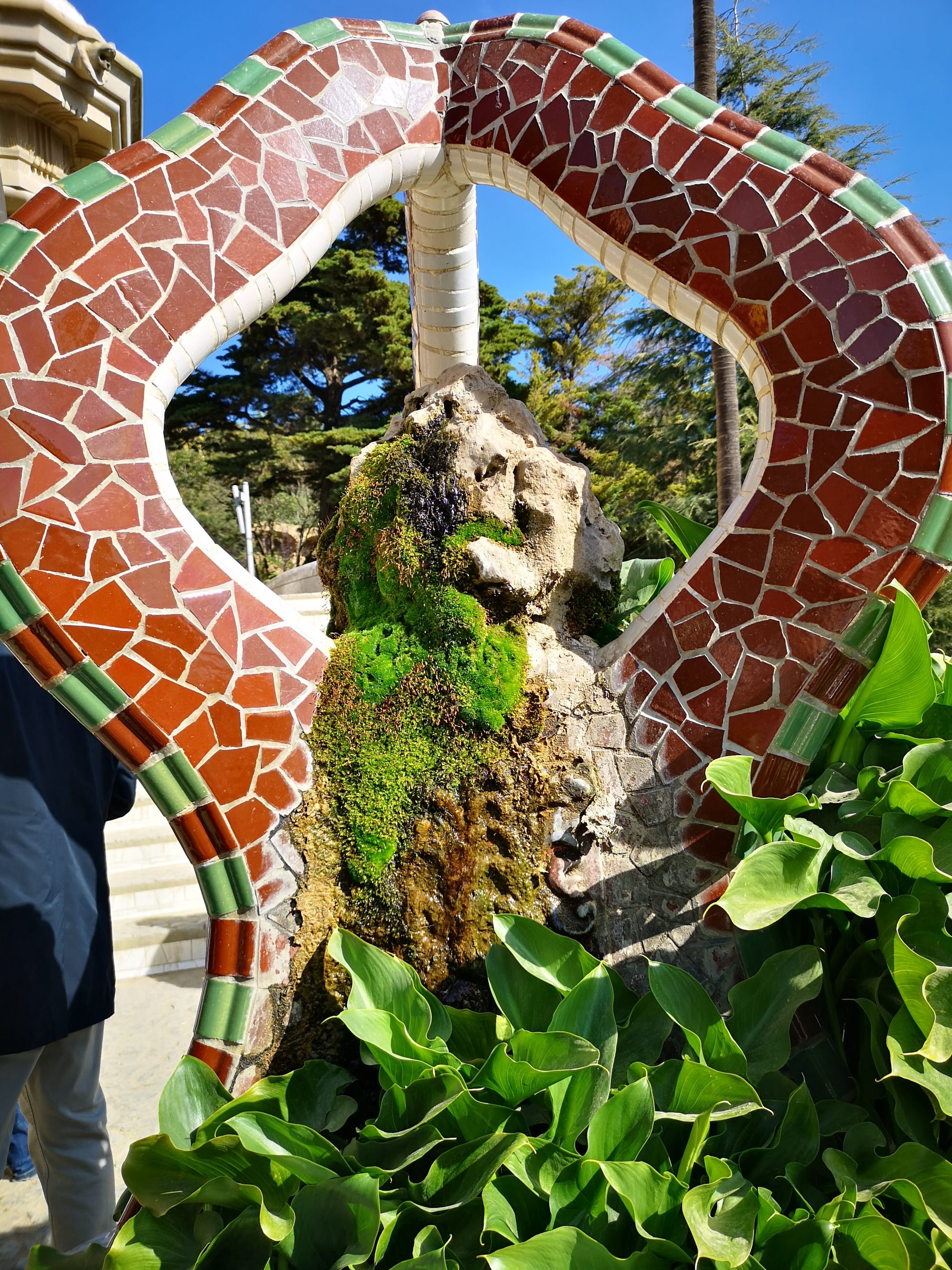 Estructura de mosaico trencadís que enmarca una fuente de roca cubierta de musgo.