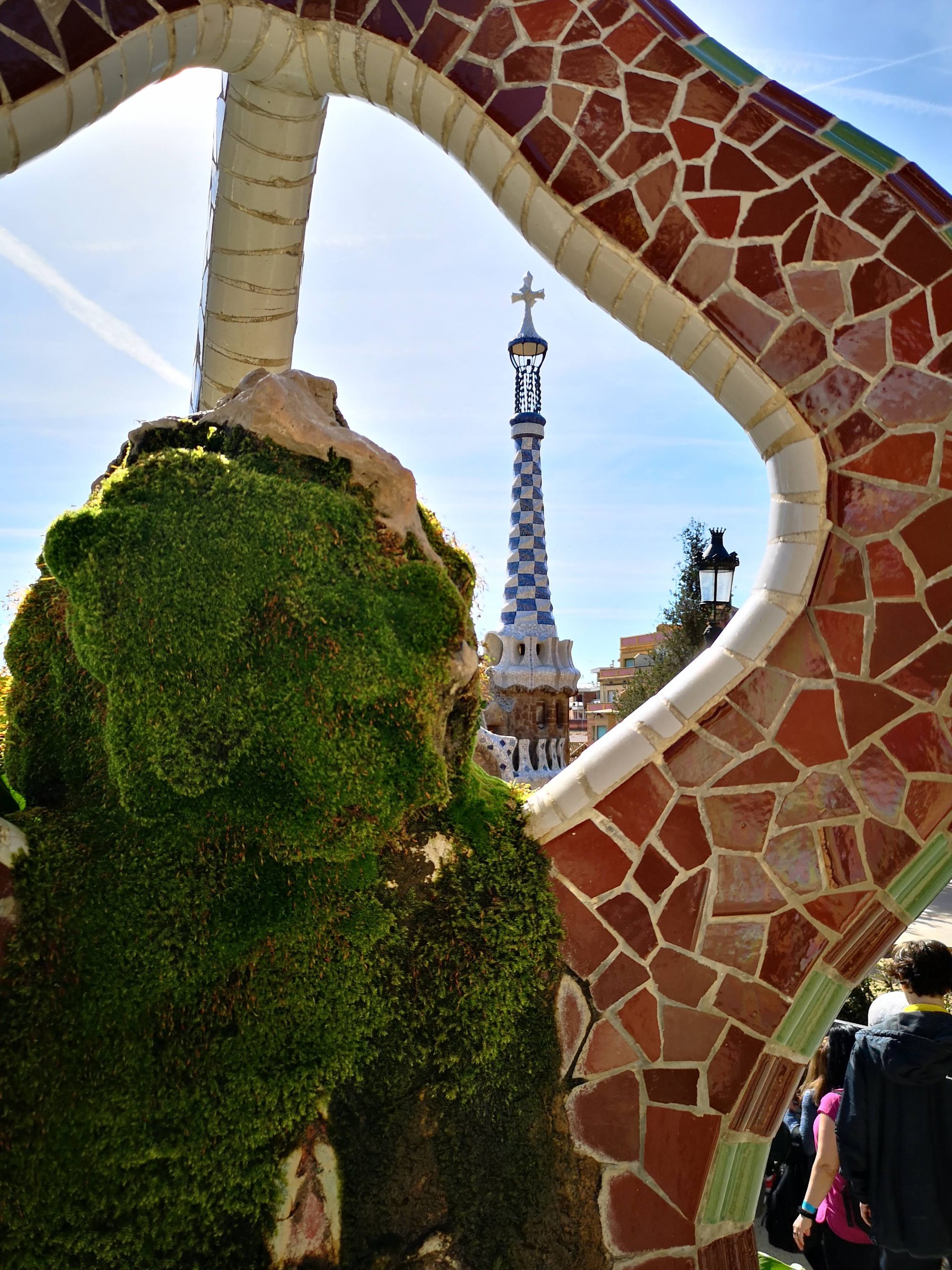 Torre del Park Güell enmarcada por un arco curvo de mosaicos trencadís y musgo.