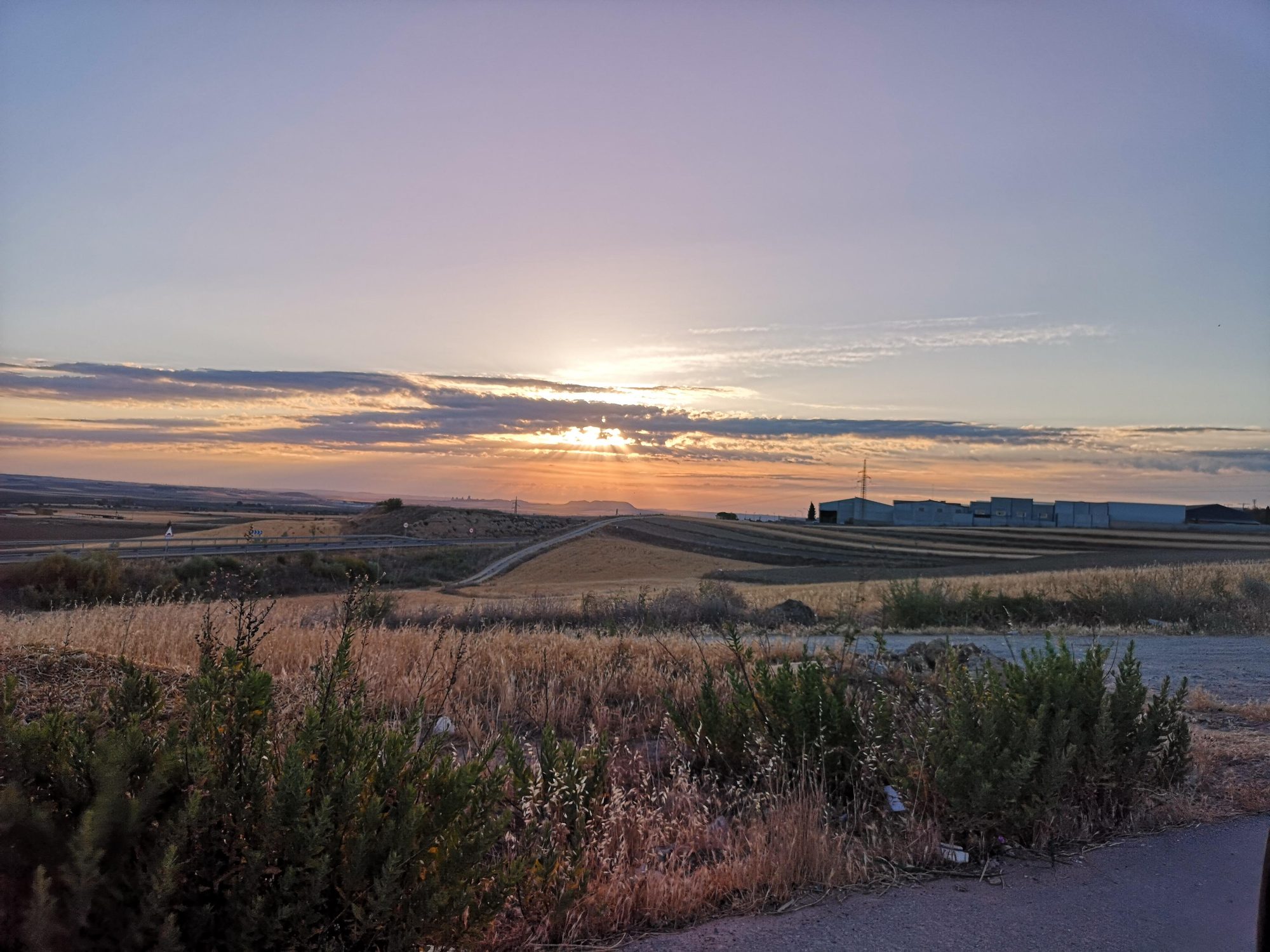 Atardecer con rayos de sol entre nubes sobre campos rurales y colinas.