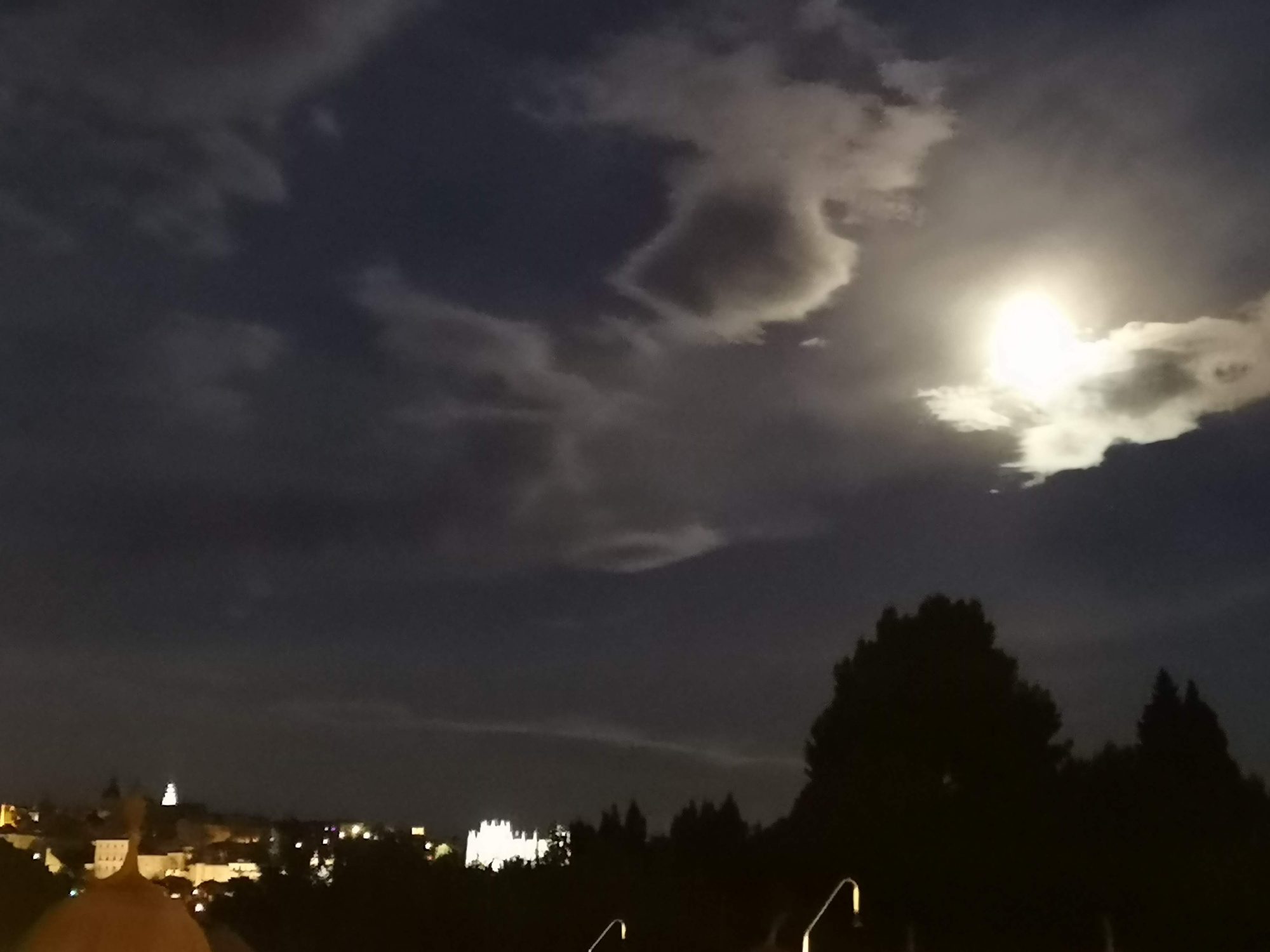Cielo nocturno con luna brillante entre nubes sobre la ciudad iluminada de Toledo.