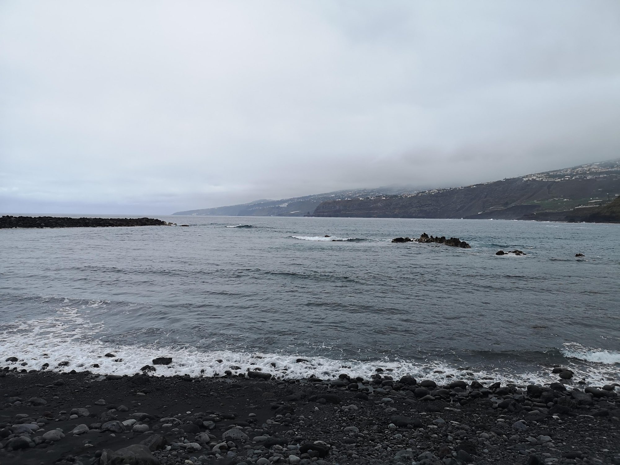 Playa de arena negra y cantos rodados con acantilados bajo un cielo nublado.