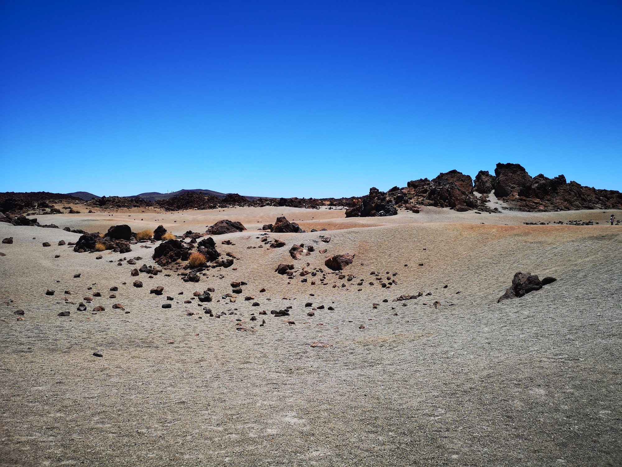 Paisaje volcánico árido con rocas oscuras y arena bajo un cielo azul despejado.
