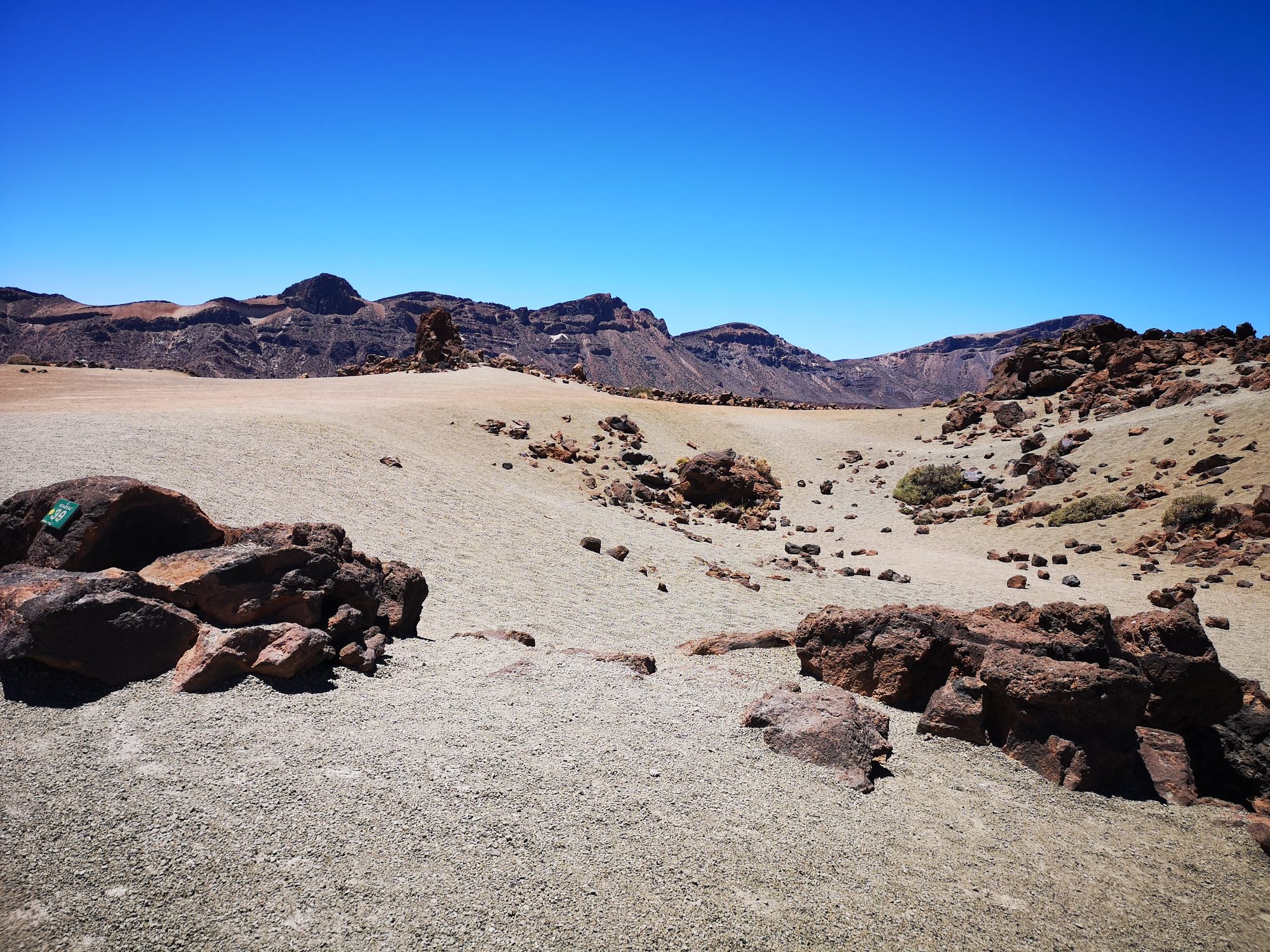 Paisaje árido y volcánico con rocas oscuras y montañas bajo un cielo azul despejado.