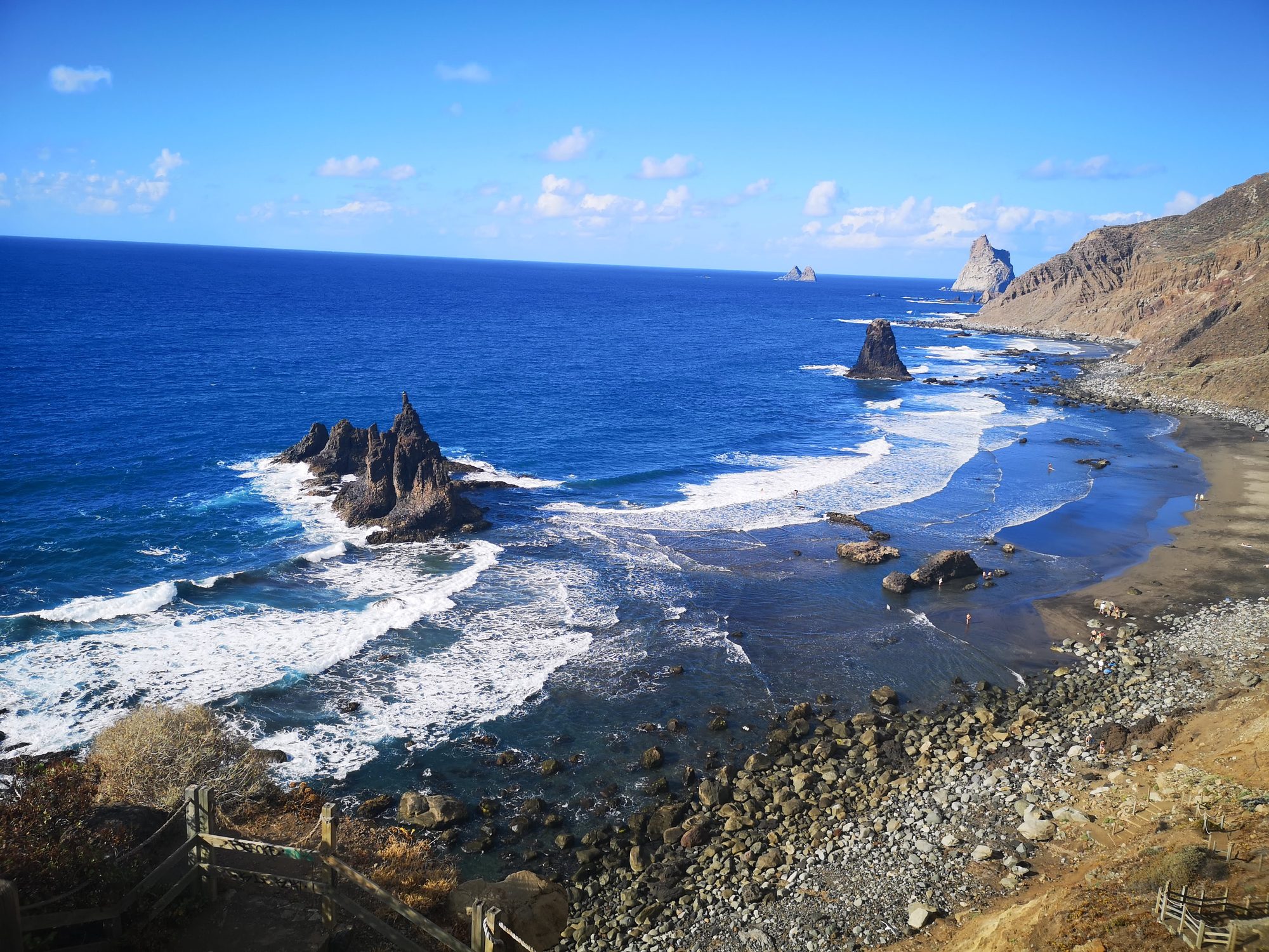Playa volcánica de arena negra con imponentes formaciones rocosas emergiendo del mar azul.