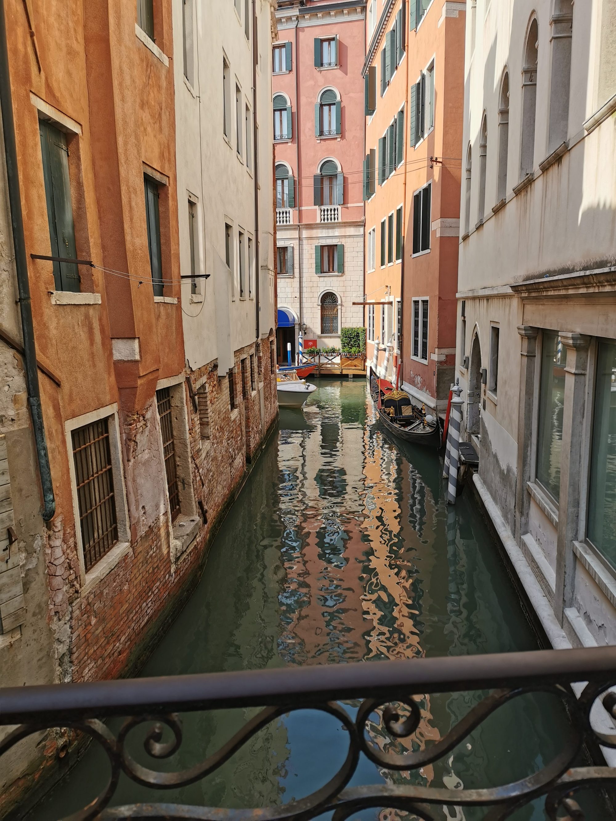 Canal estrecho de Venecia con edificios coloridos, reflejos en el agua y una góndola amarrada.