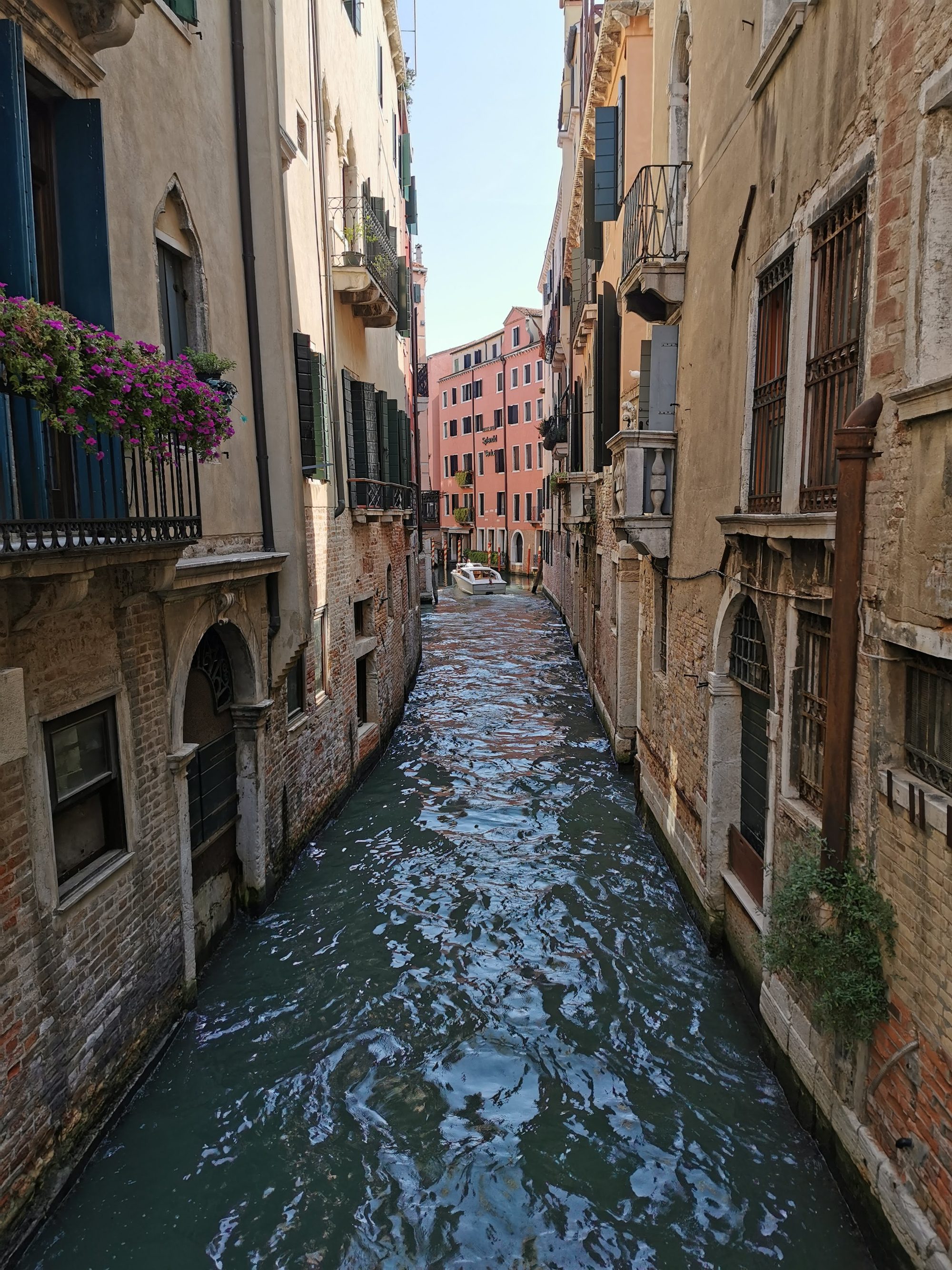 Estrecho canal de Venecia flanqueado por edificios históricos con balcones floridos y una lancha.