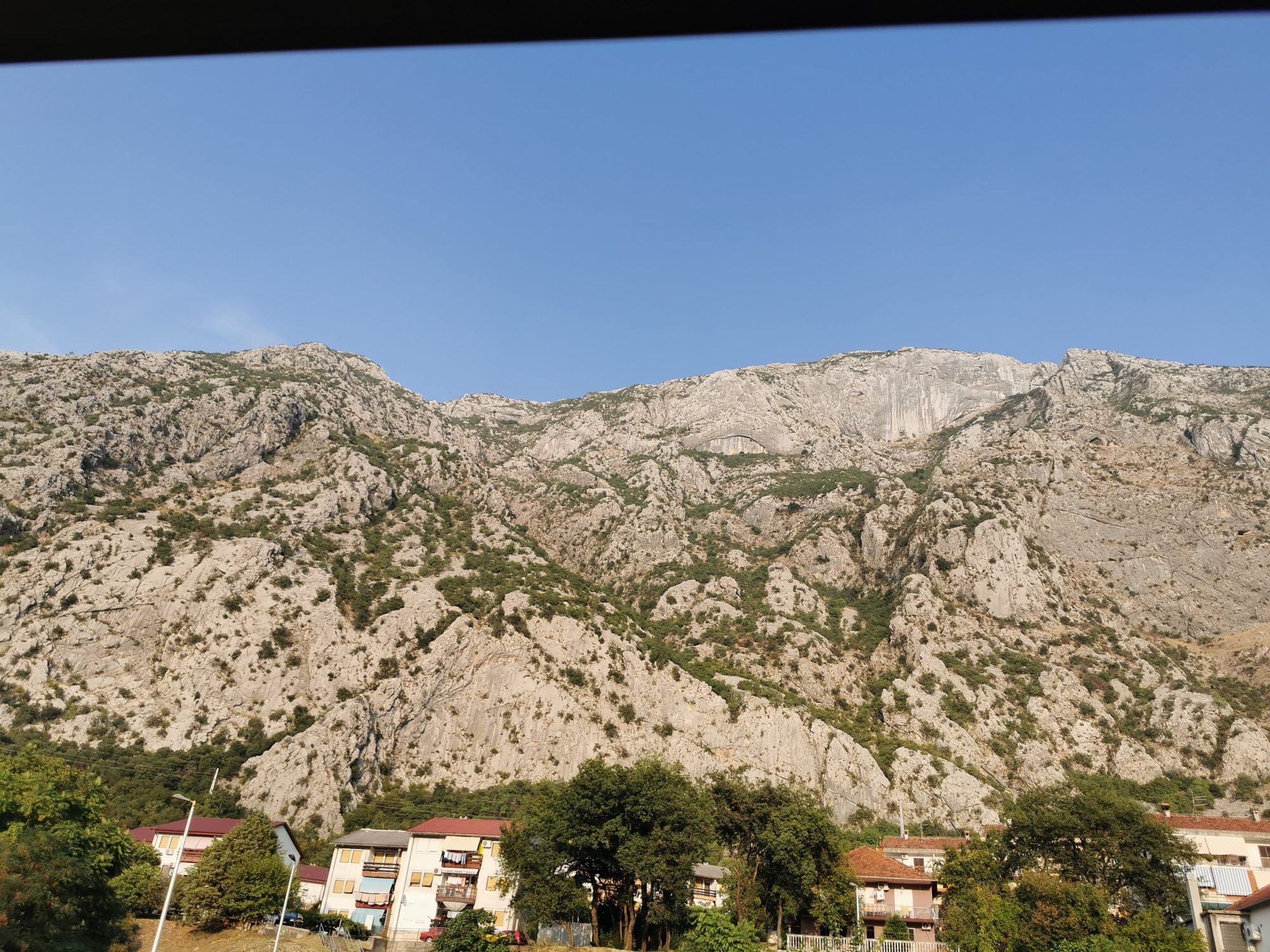 Imponentes montañas rocosas elevándose sobre casas y árboles en Dobrota bajo cielo despejado.