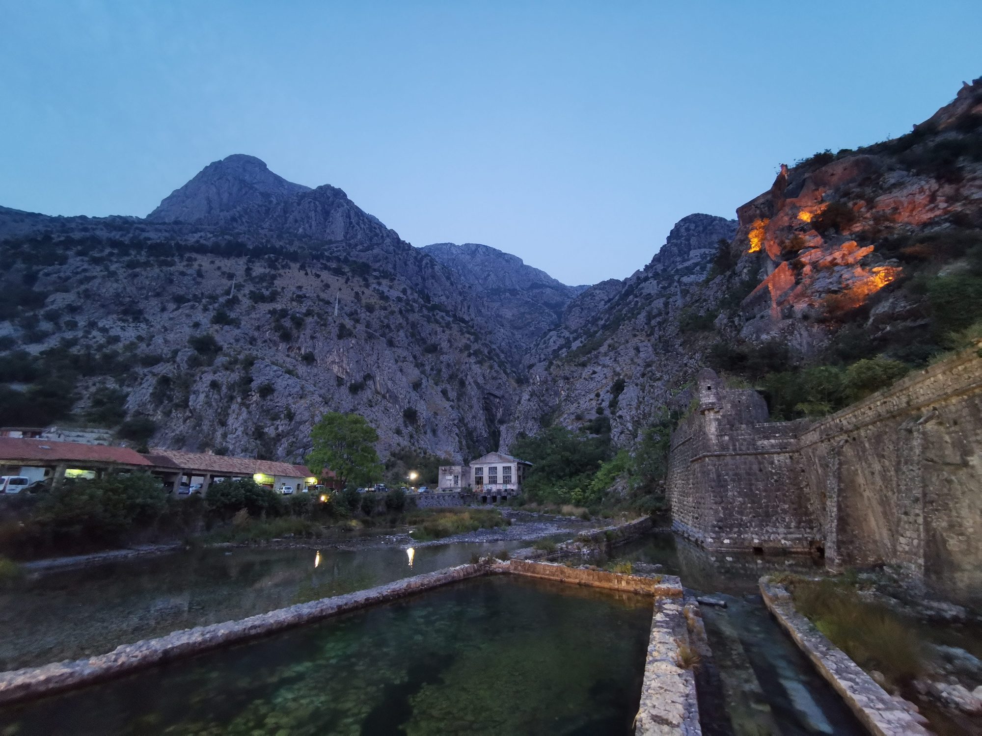 Murallas históricas de piedra y canales de agua frente a escarpadas montañas al anochecer.