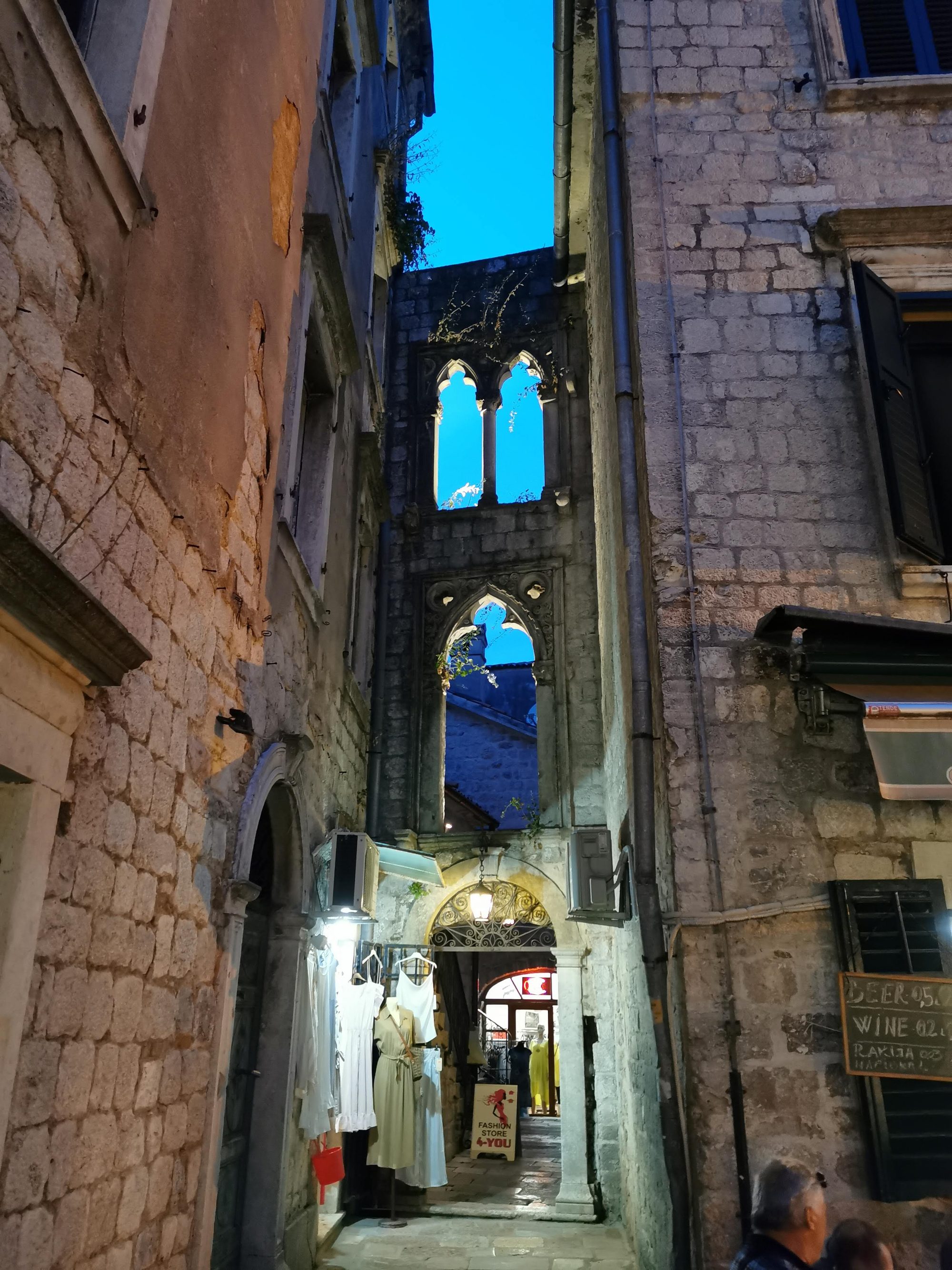 Callejón medieval de piedra con ventanales góticos abiertos al cielo azul en Kotor.