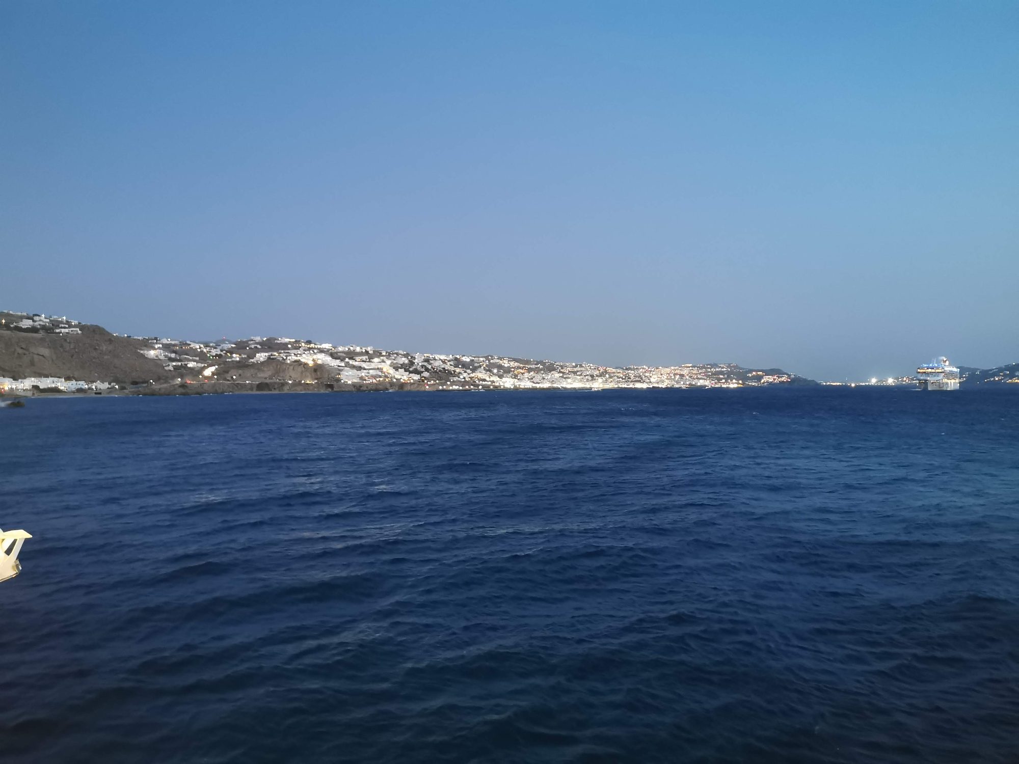 Costa iluminada de Miconos al anochecer con el mar y un crucero navegando.