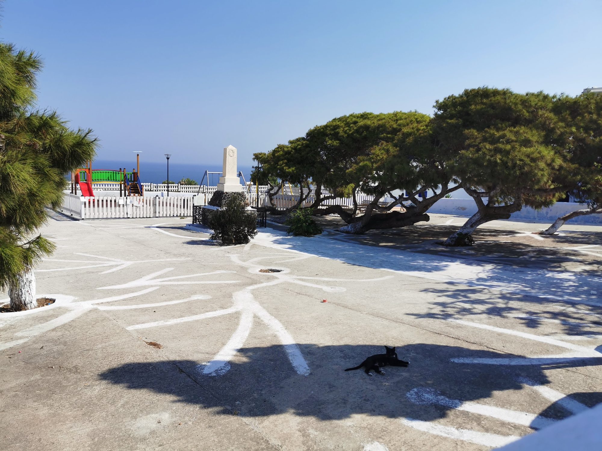 Plaza con suelo pintado, árboles frondosos, un monumento y un gato a la sombra.