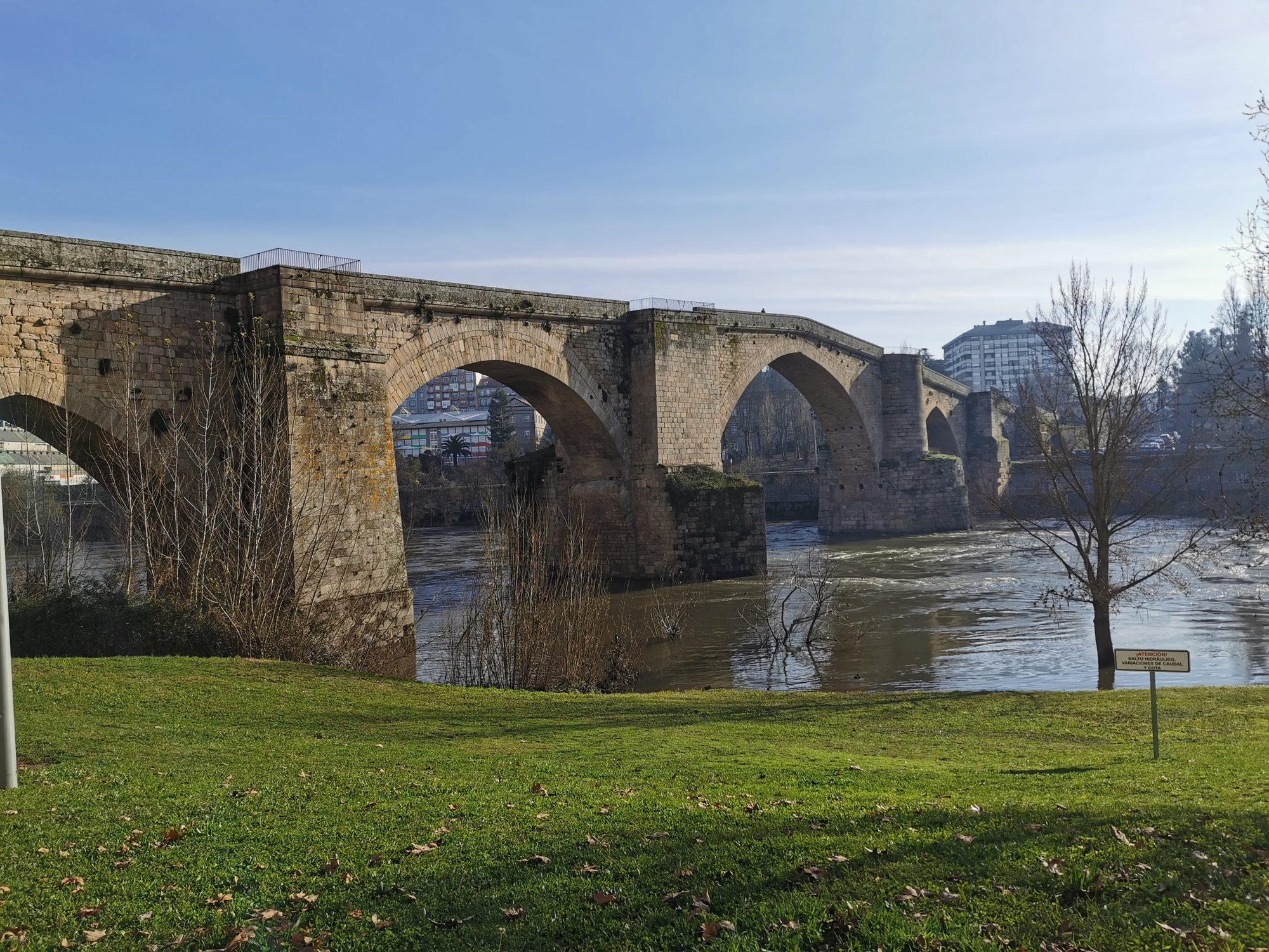 Puente romano de piedra con arcos sobre el río Miño junto a una orilla verde.