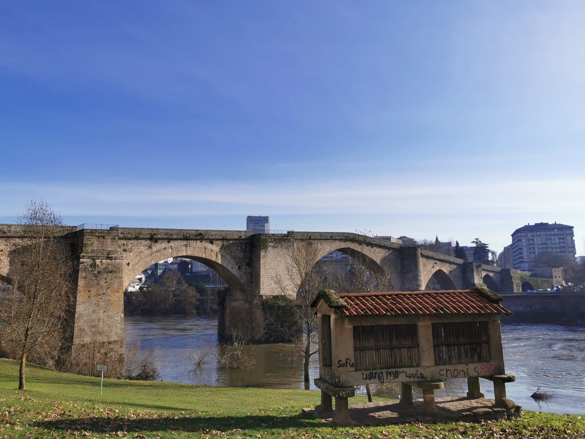 Hórreo tradicional gallego frente al Puente Romano de Ourense sobre el río Miño.