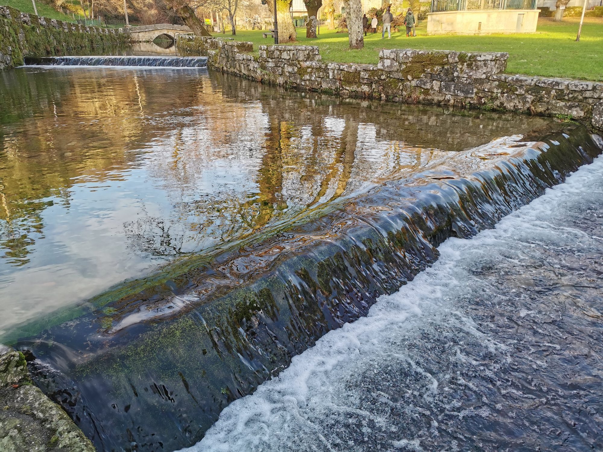 Pequeña presa en un río con reflejos de árboles y parque al fondo.