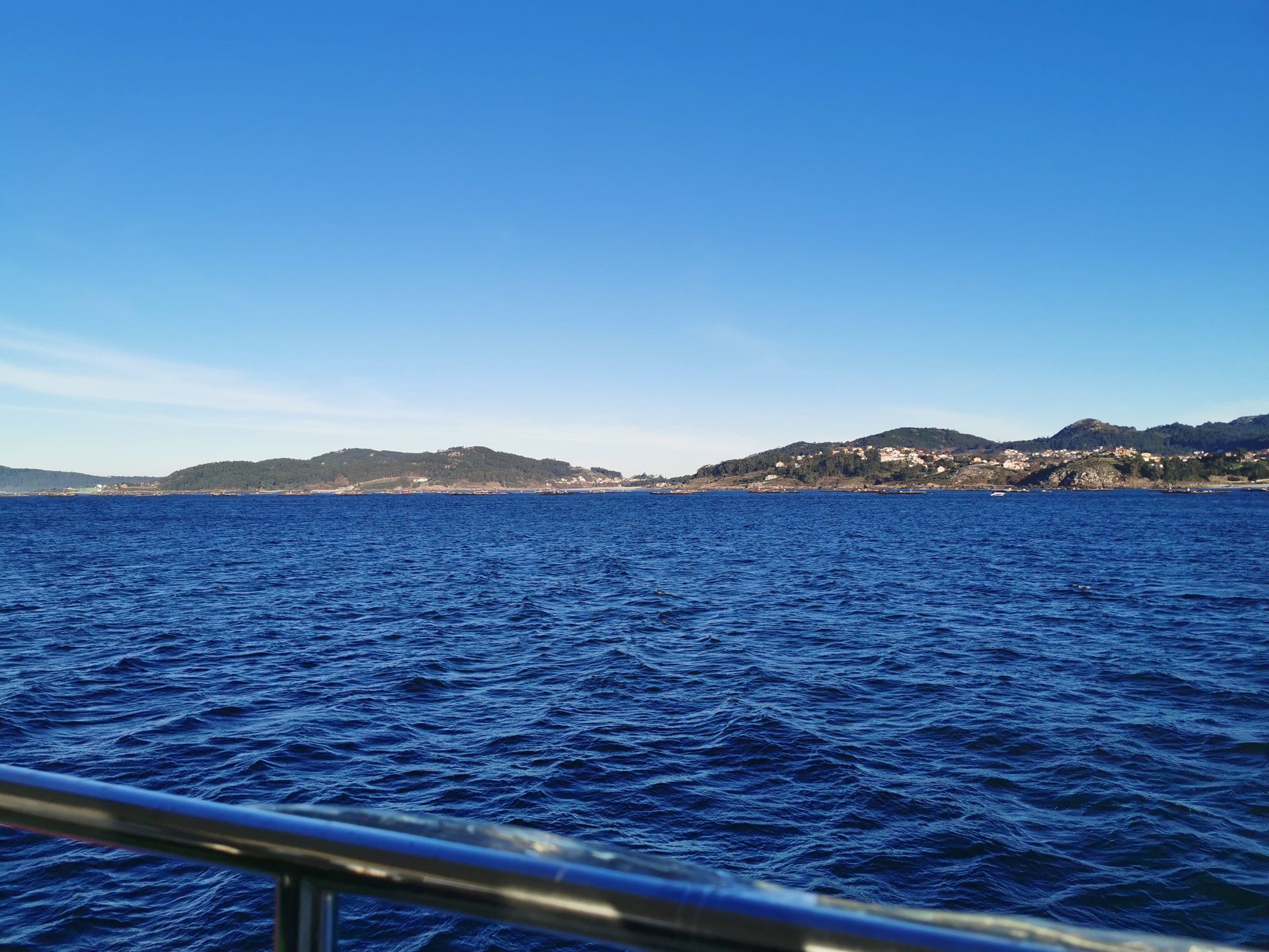 Costa montañosa y bateas vistas desde un barco navegando por el mar azul.