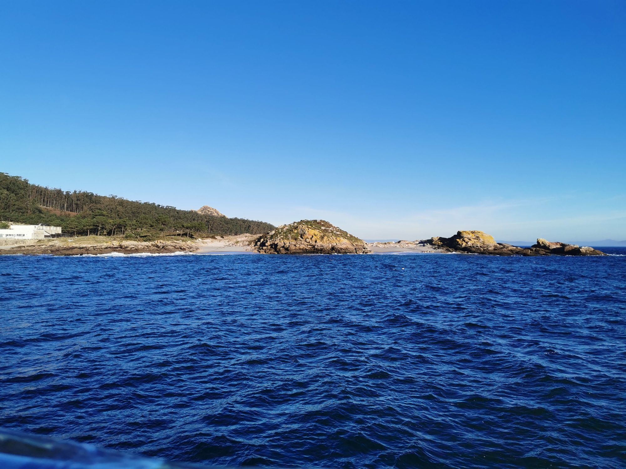 Costa rocosa con pequeña playa y pinares vista desde el mar azul intenso.
