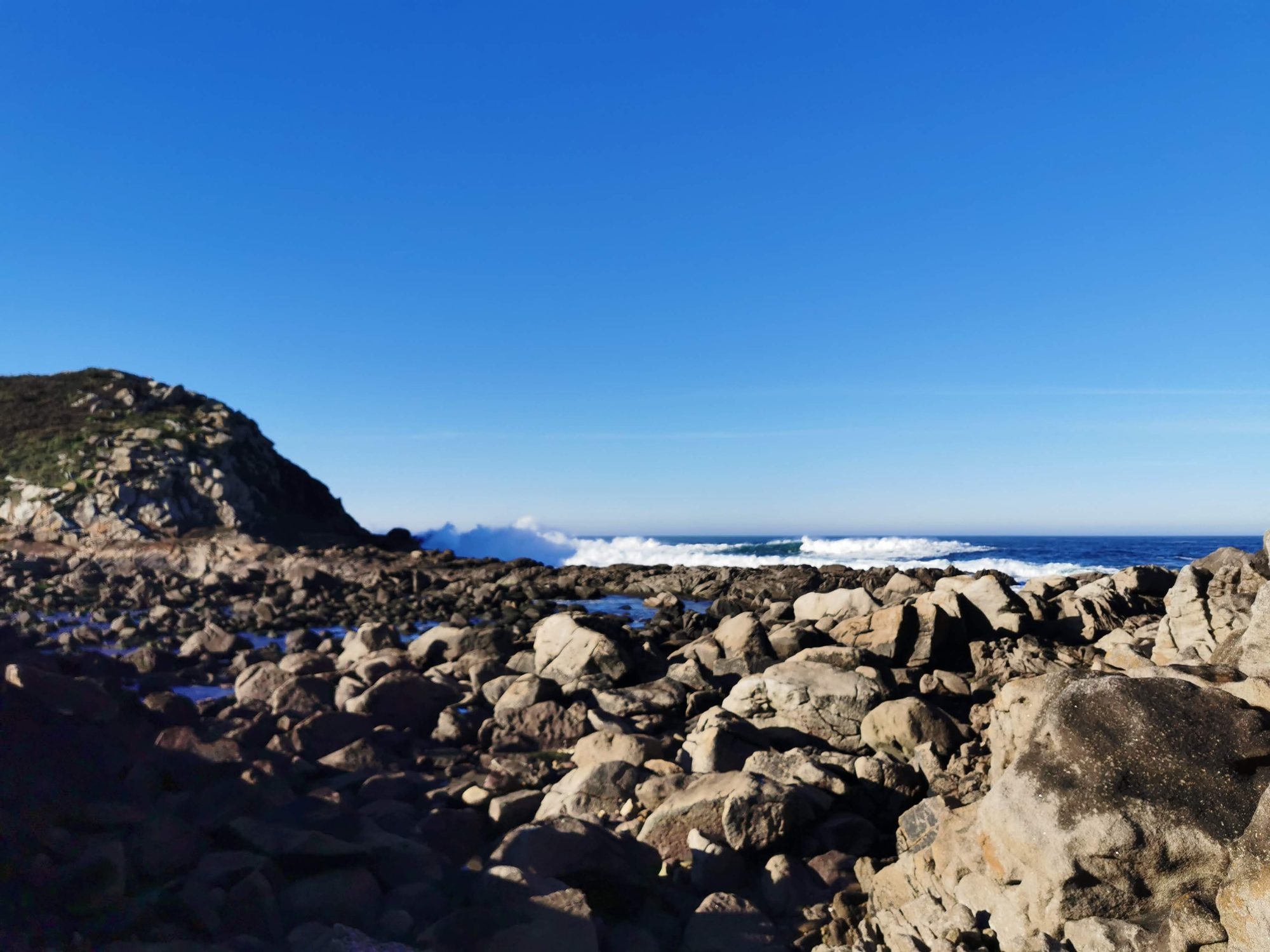 Costa rocosa junto al mar con olas rompiendo bajo un cielo azul despejado.