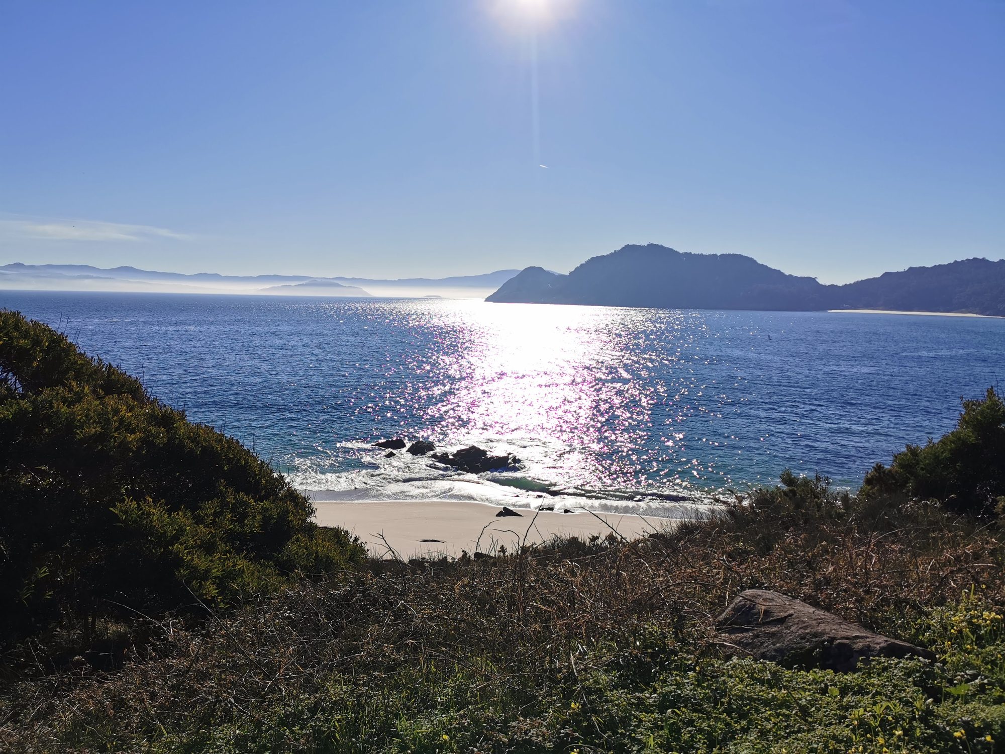 Destello de sol sobre el mar y pequeña playa rodeada de colinas y vegetación.