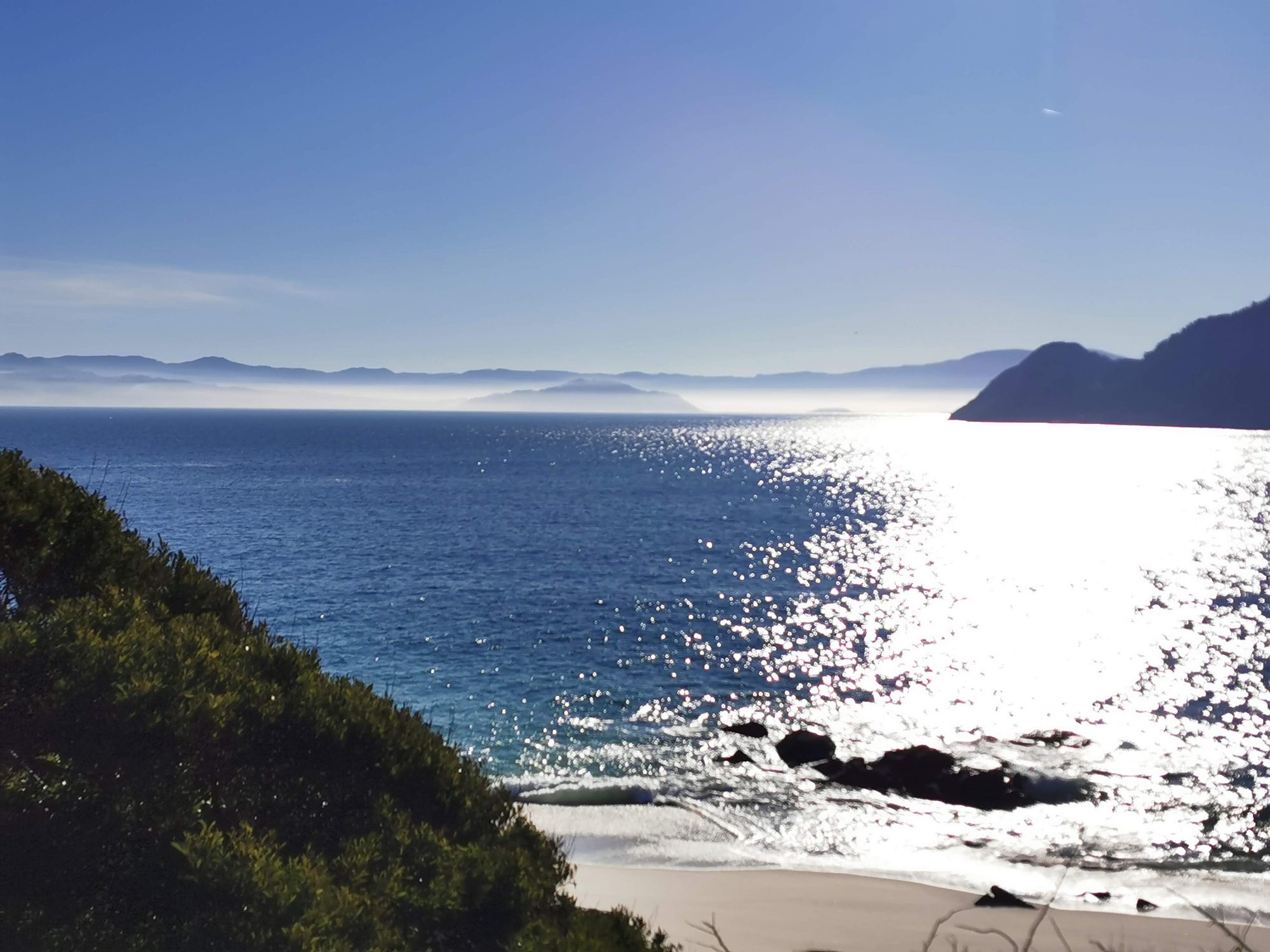 Playa costera y mar resplandeciente bajo el sol con colinas y niebla al fondo.