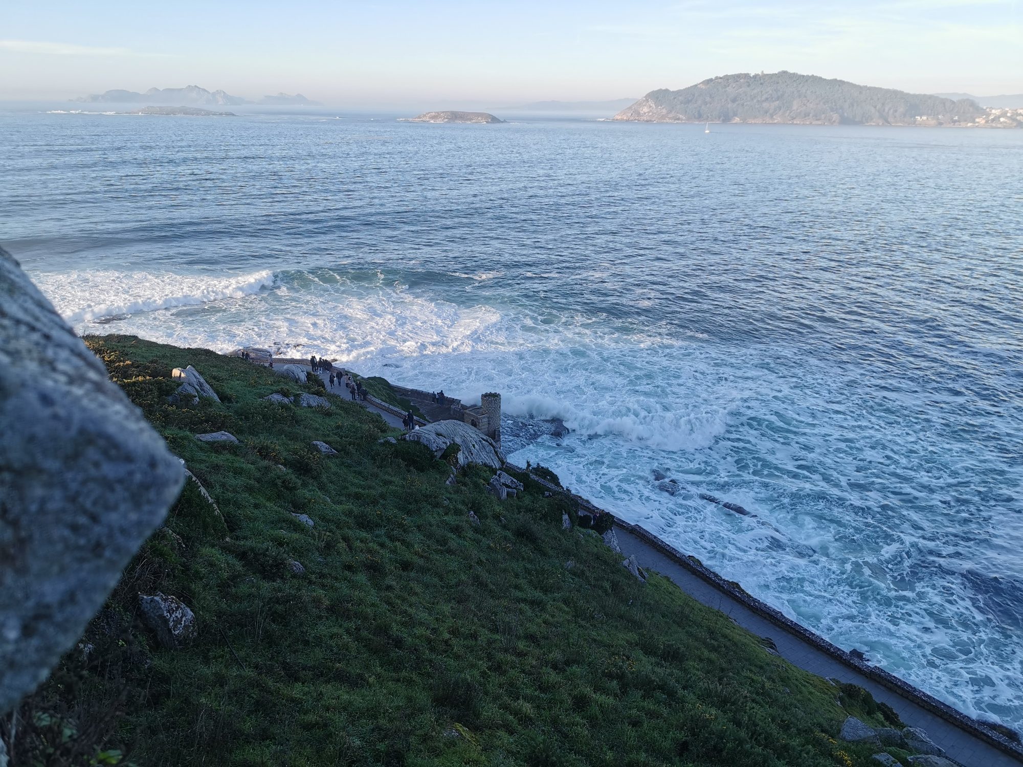 Paseo costero y olas rompiendo junto a la ladera verde con islas al fondo.