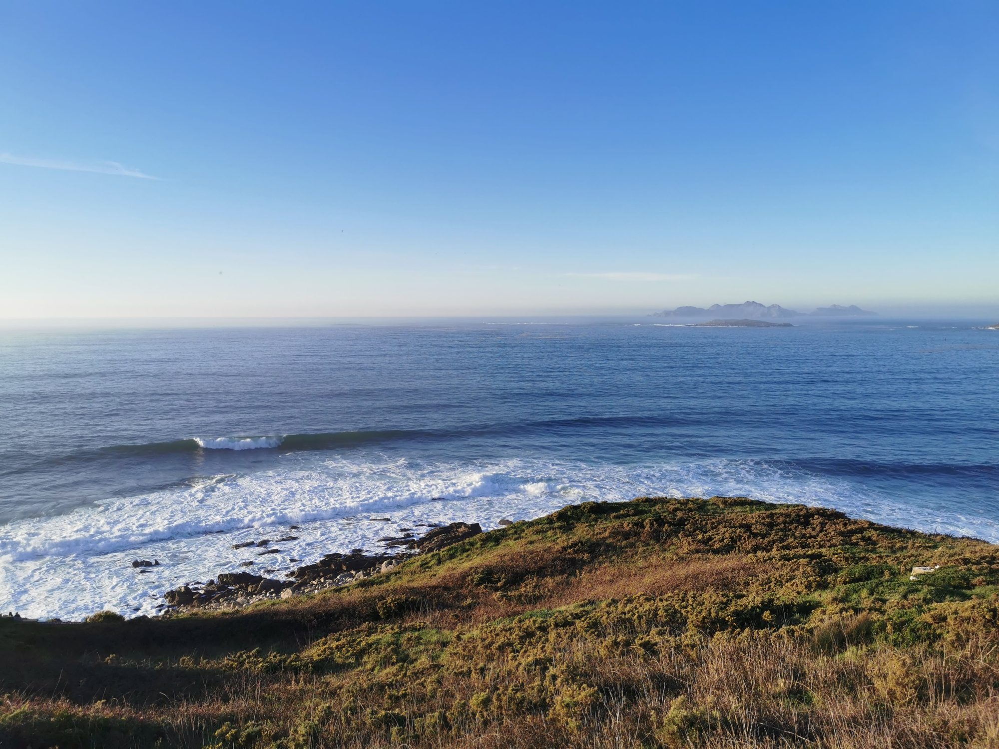 Olas del océano Atlántico rompiendo en la costa rocosa y vegetación en Baiona.