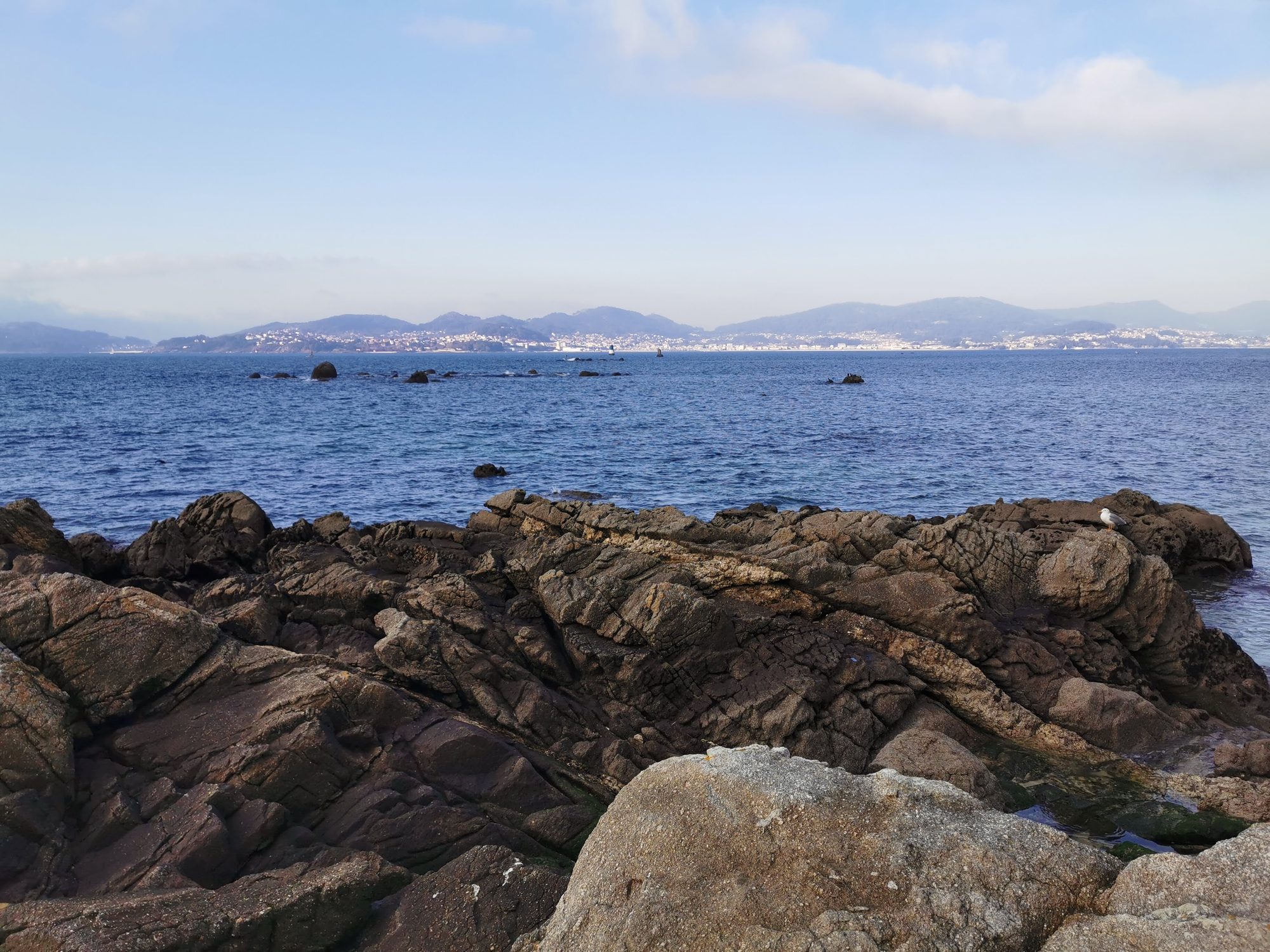 Costa rocosa frente al mar azul con montañas y pueblos en el horizonte.