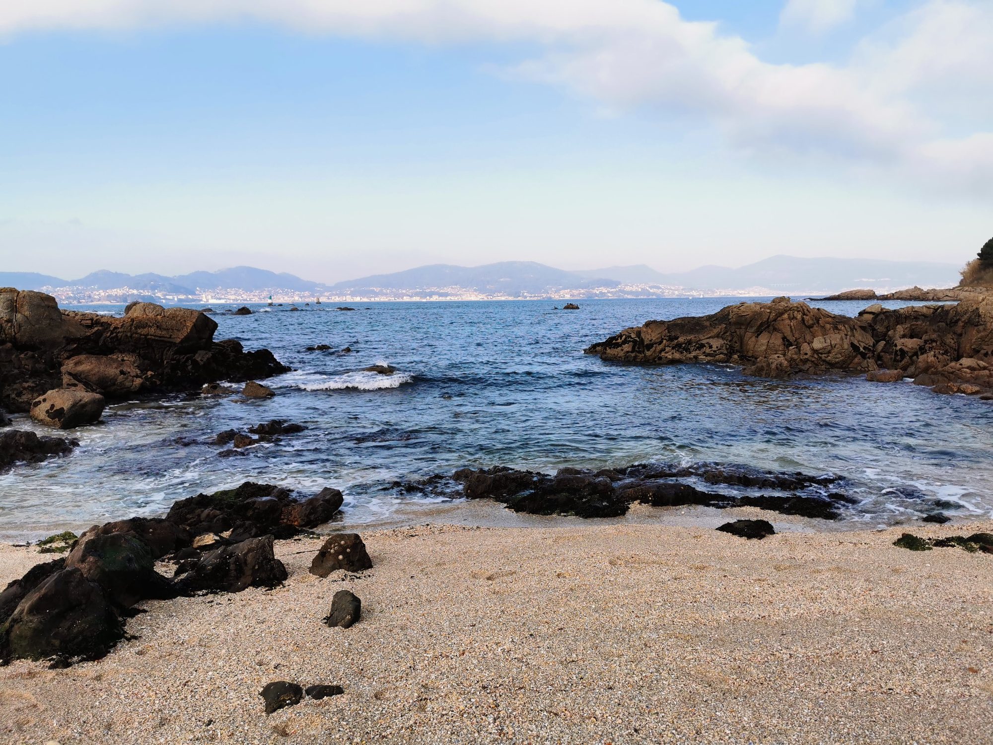 Caleta rocosa con arena gruesa y vistas a la ría de Vigo bajo cielo azul.