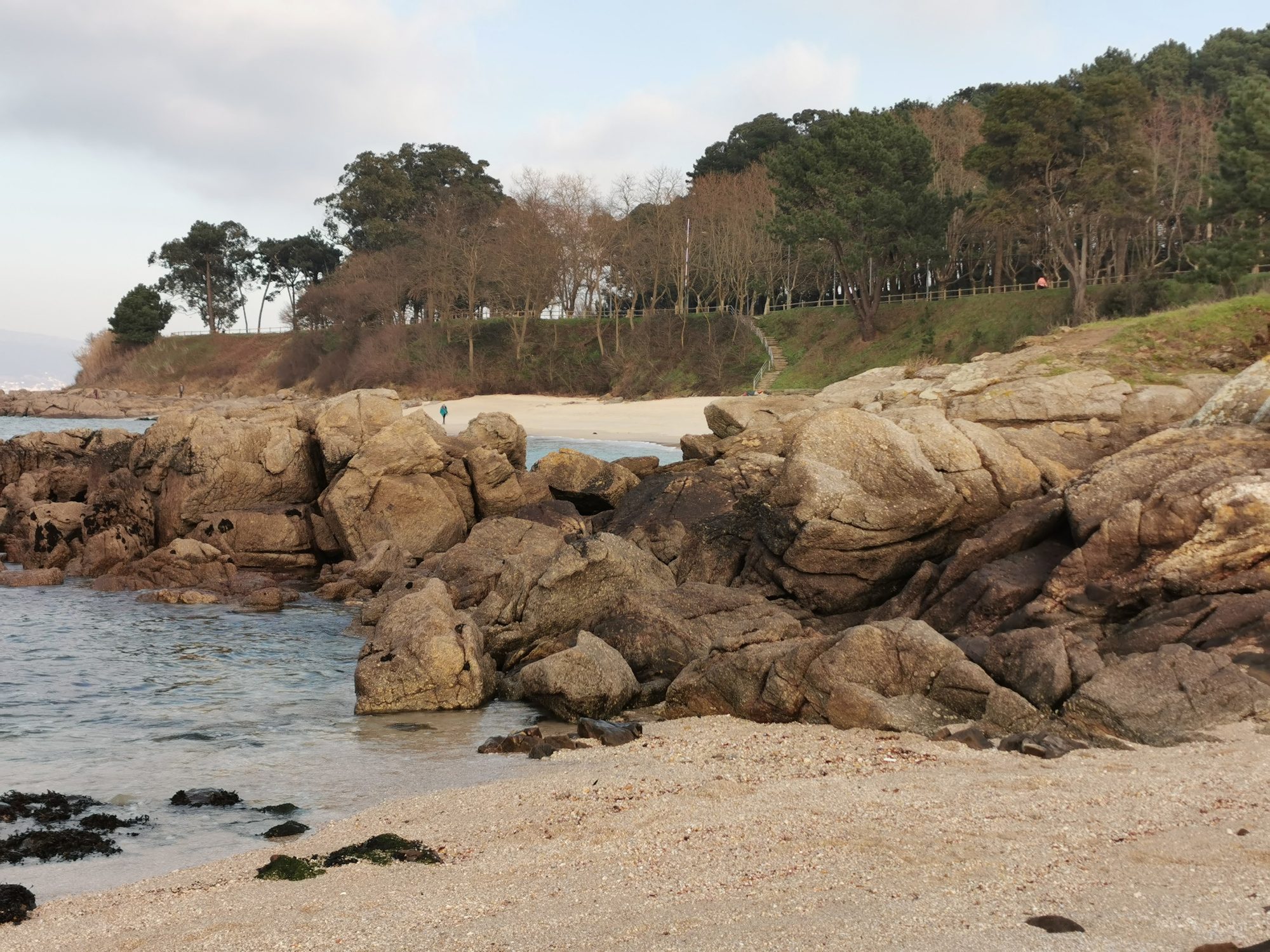 Grandes rocas graníticas en la arena de una cala costera con bosque al fondo.