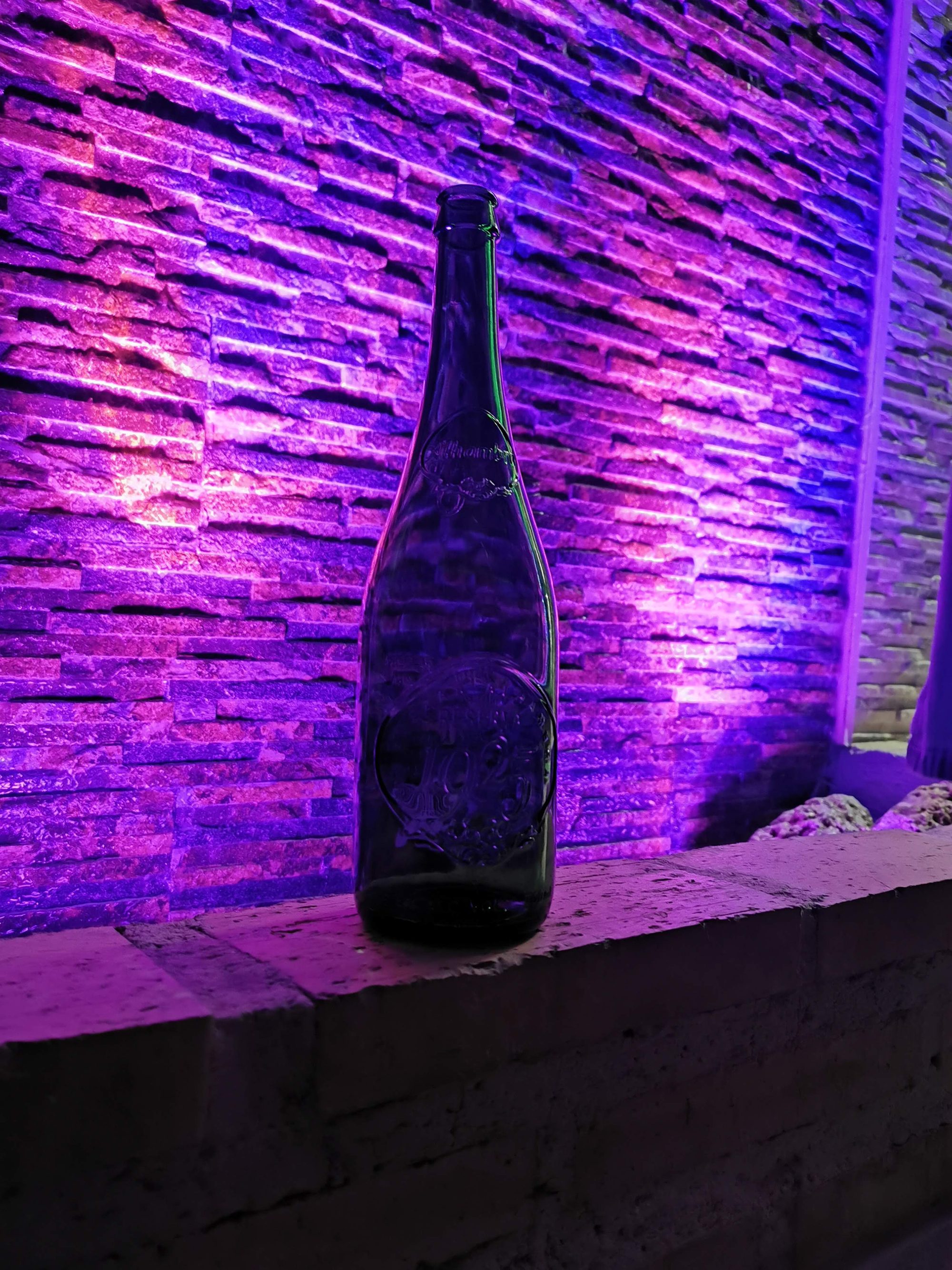Botella de vidrio verde sobre un muro iluminado con luz violeta de fondo.