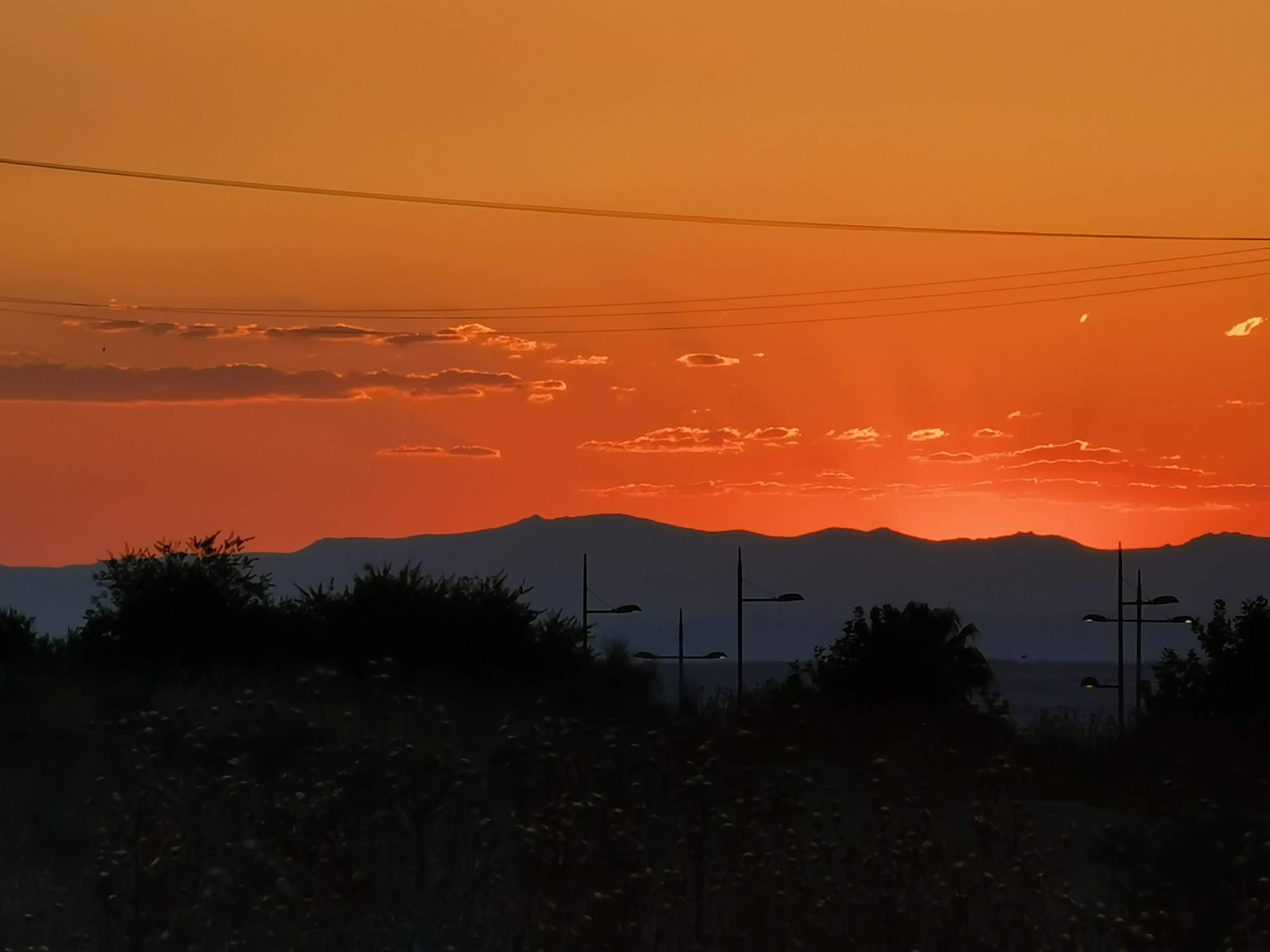 Cielo anaranjado de atardecer sobre la silueta de montañas, farolas y árboles.