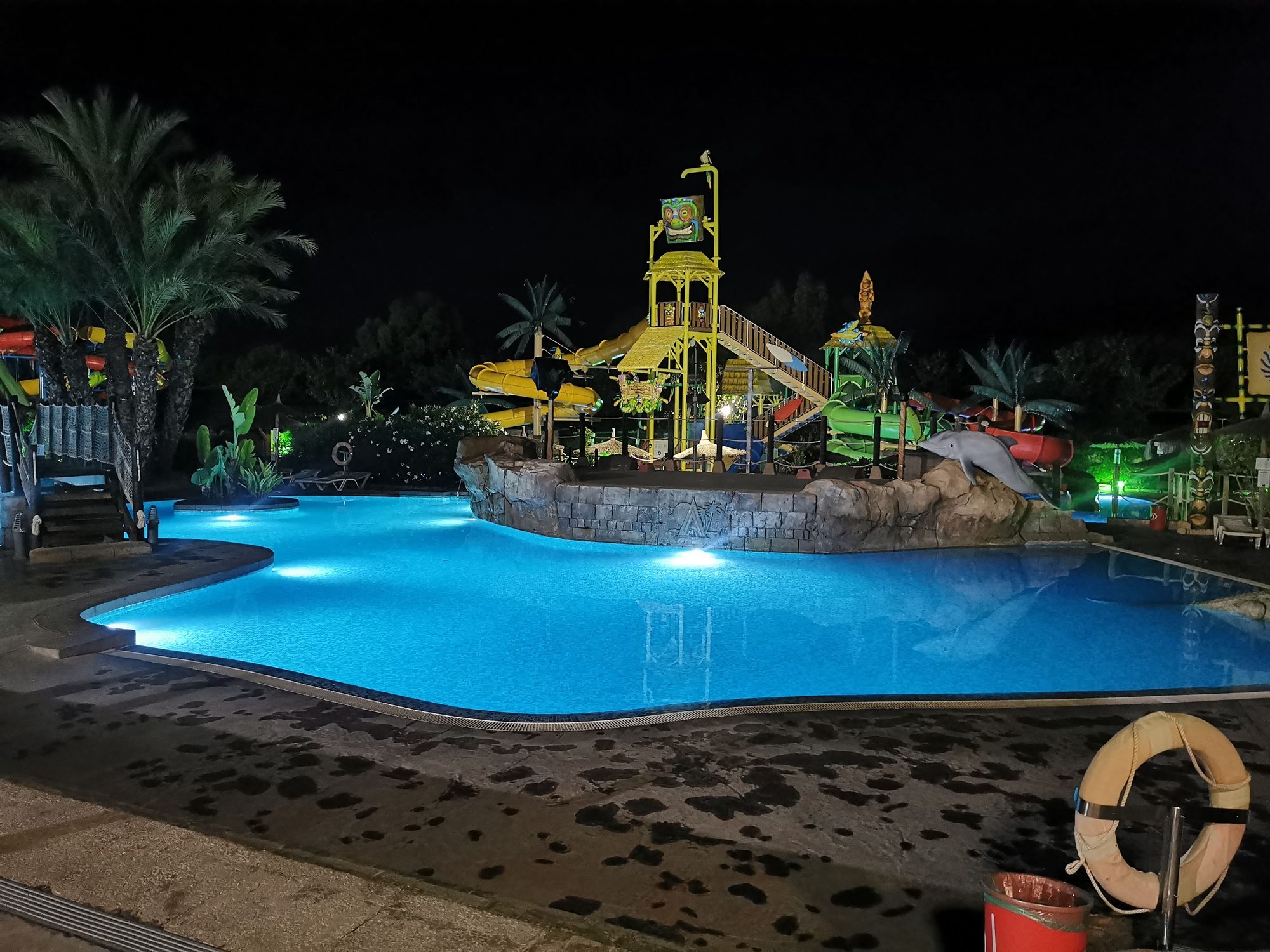 Piscina iluminada de noche con toboganes y atracciones acuáticas infantiles entre palmeras.