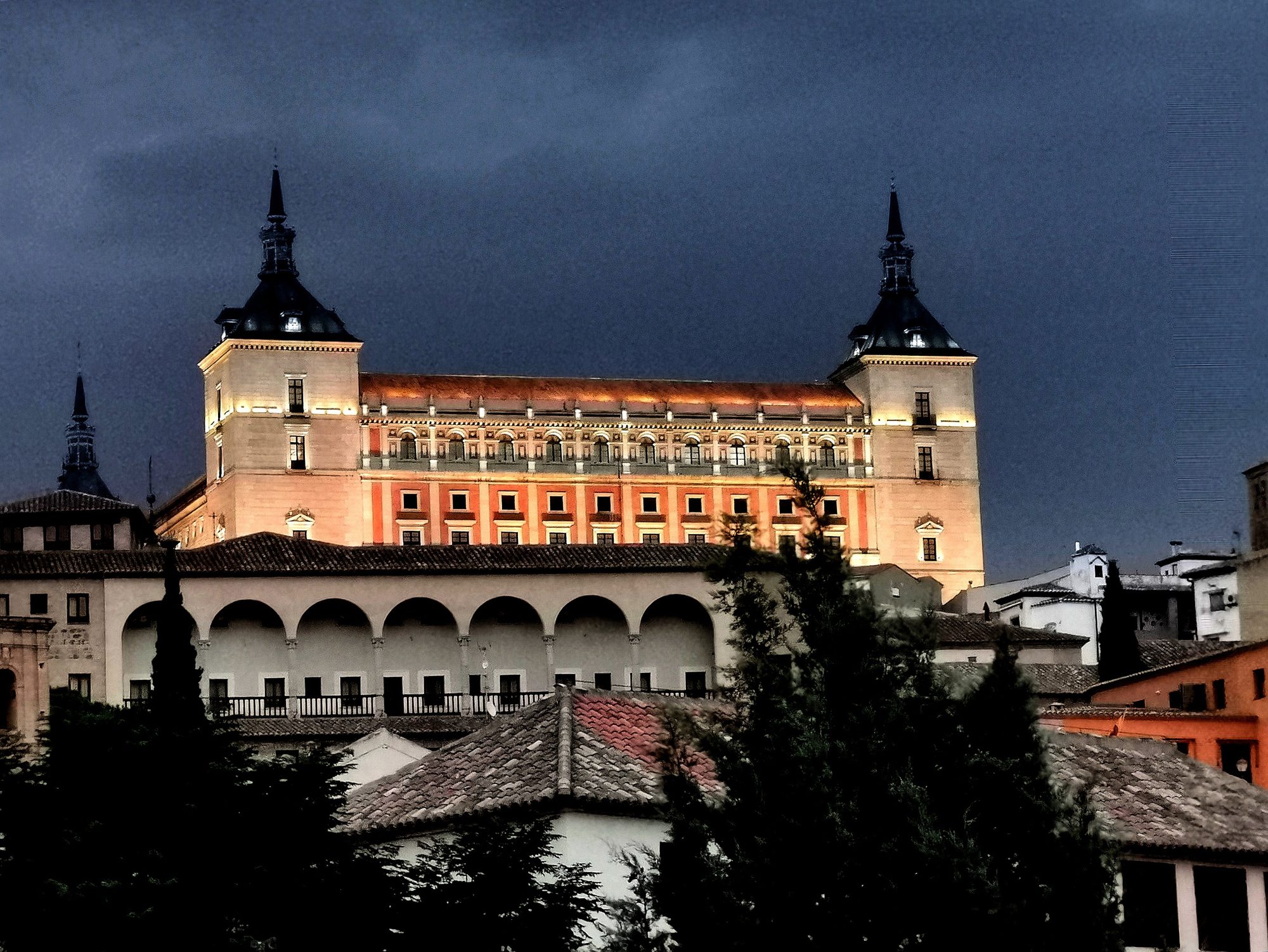 Alcázar de Toledo iluminado al anochecer sobre los tejados y arquerías de la ciudad.