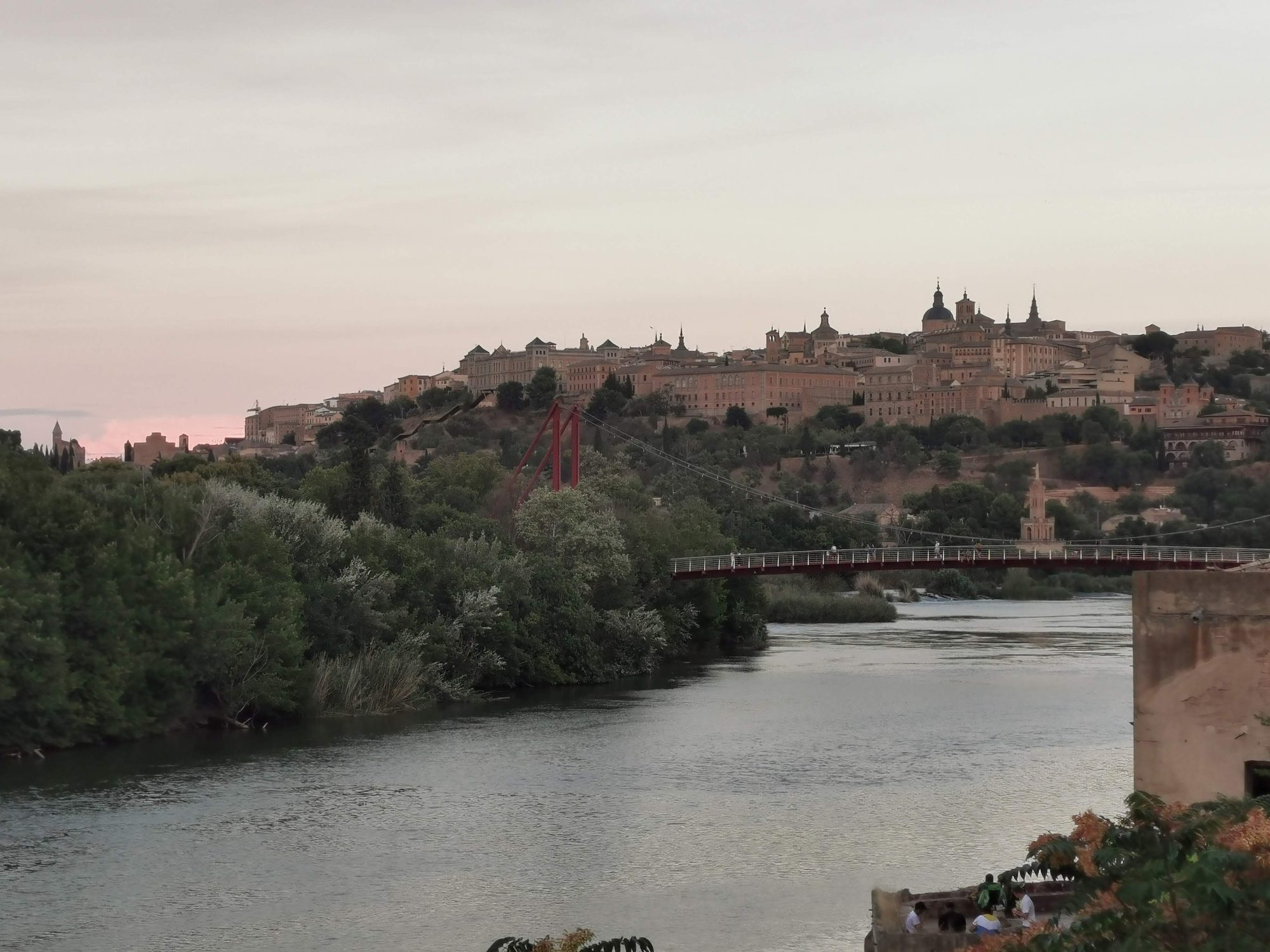 Panorámica de Toledo con el río Tajo y un puente peatonal al atardecer.