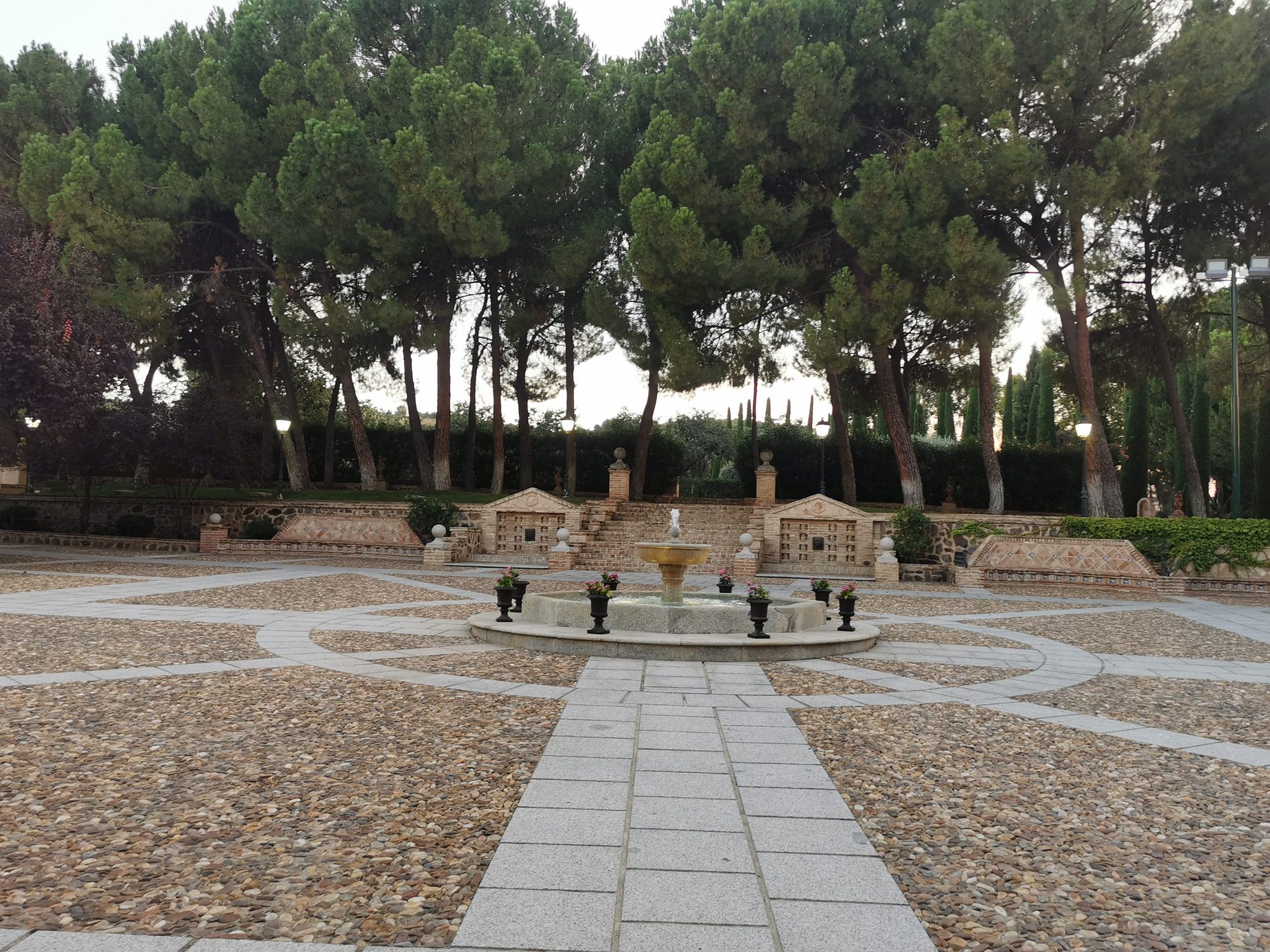 Plaza empedrada con fuente central rodeada de altos pinos y bancos decorativos de piedra.