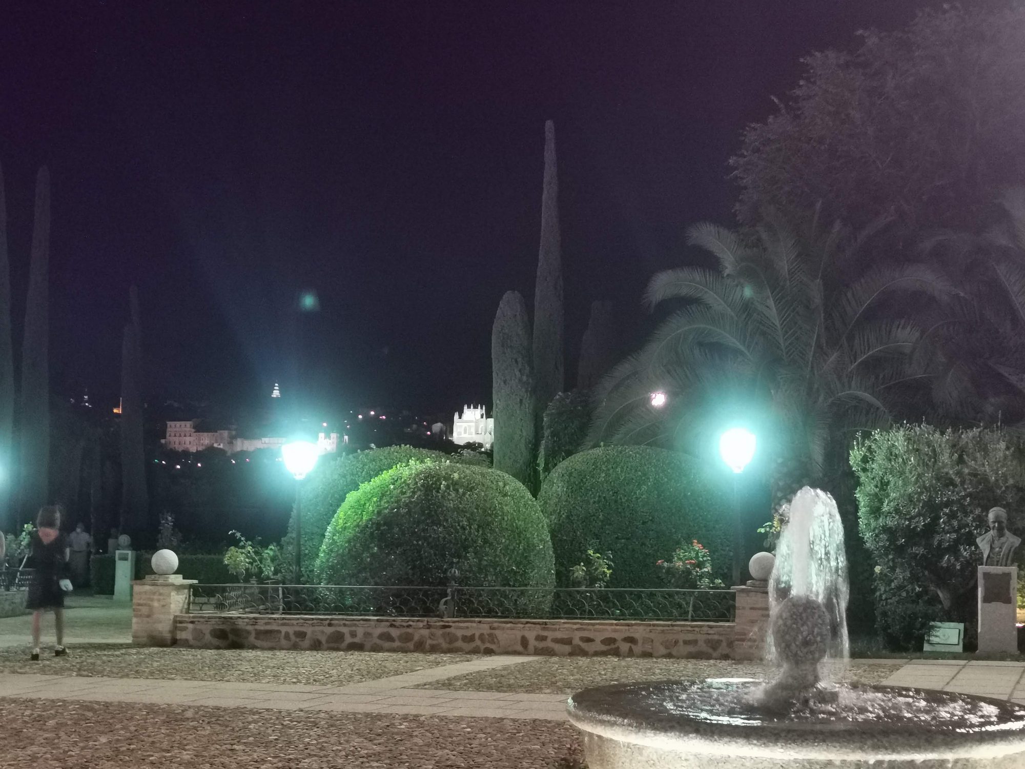 Jardines iluminados de noche con una fuente en primer plano y cipreses recortados.