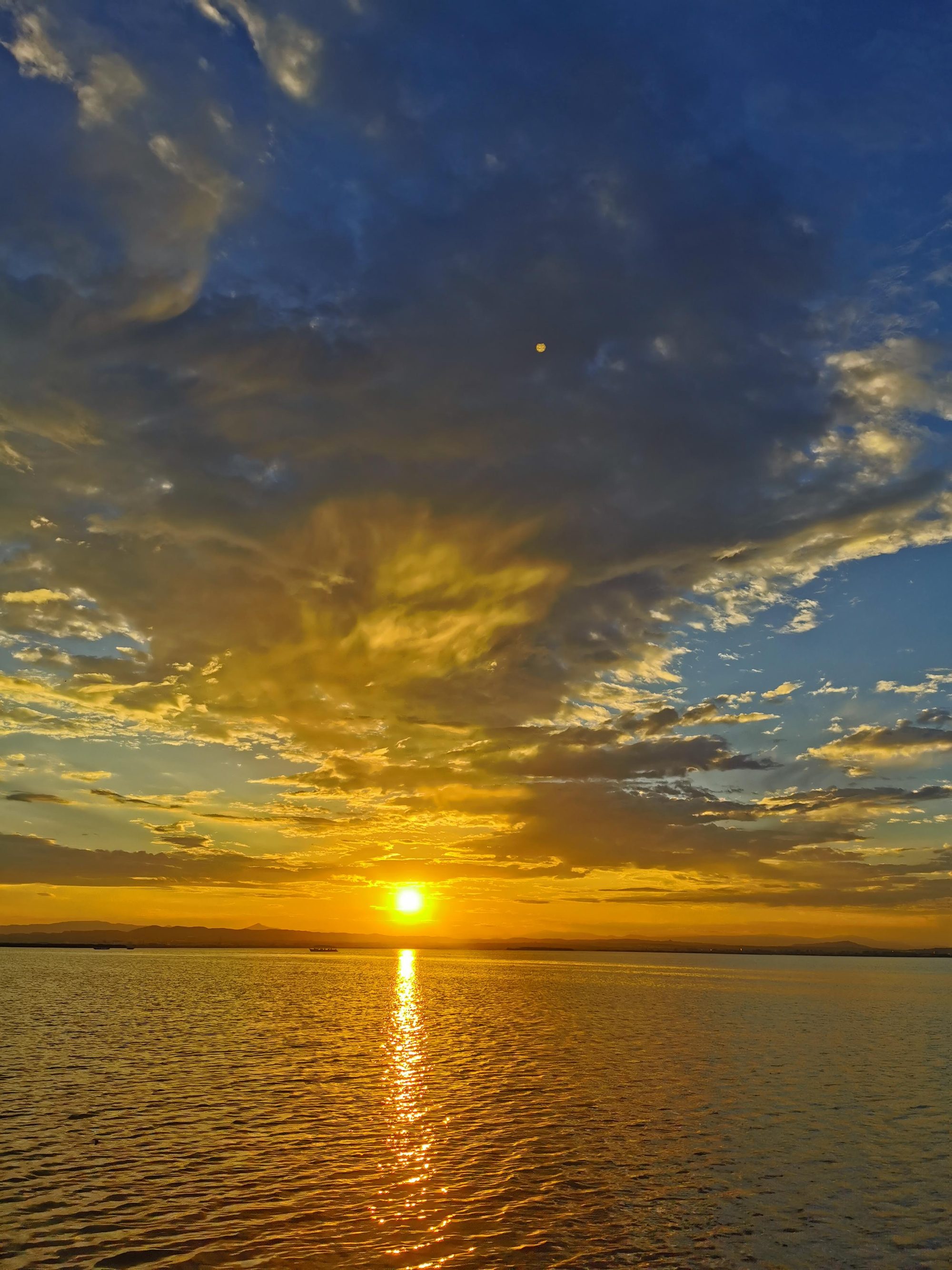 Puesta de sol dorada sobre el agua con nubes iluminadas y cielo azul.