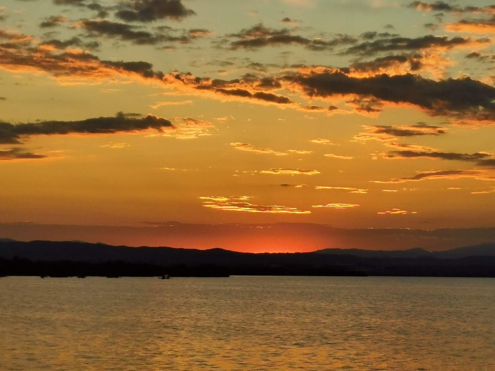 Atardecer dorado sobre el agua con siluetas de montañas y nubes iluminadas en el horizonte.