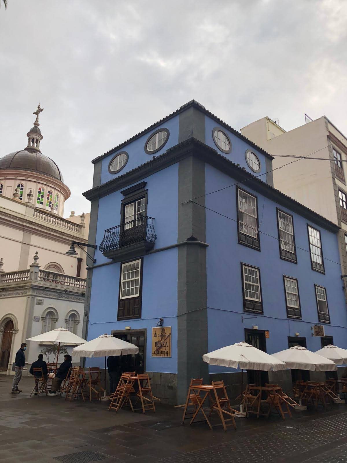Edificio azul colonial con terraza de cafetería y cúpula de iglesia al fondo.