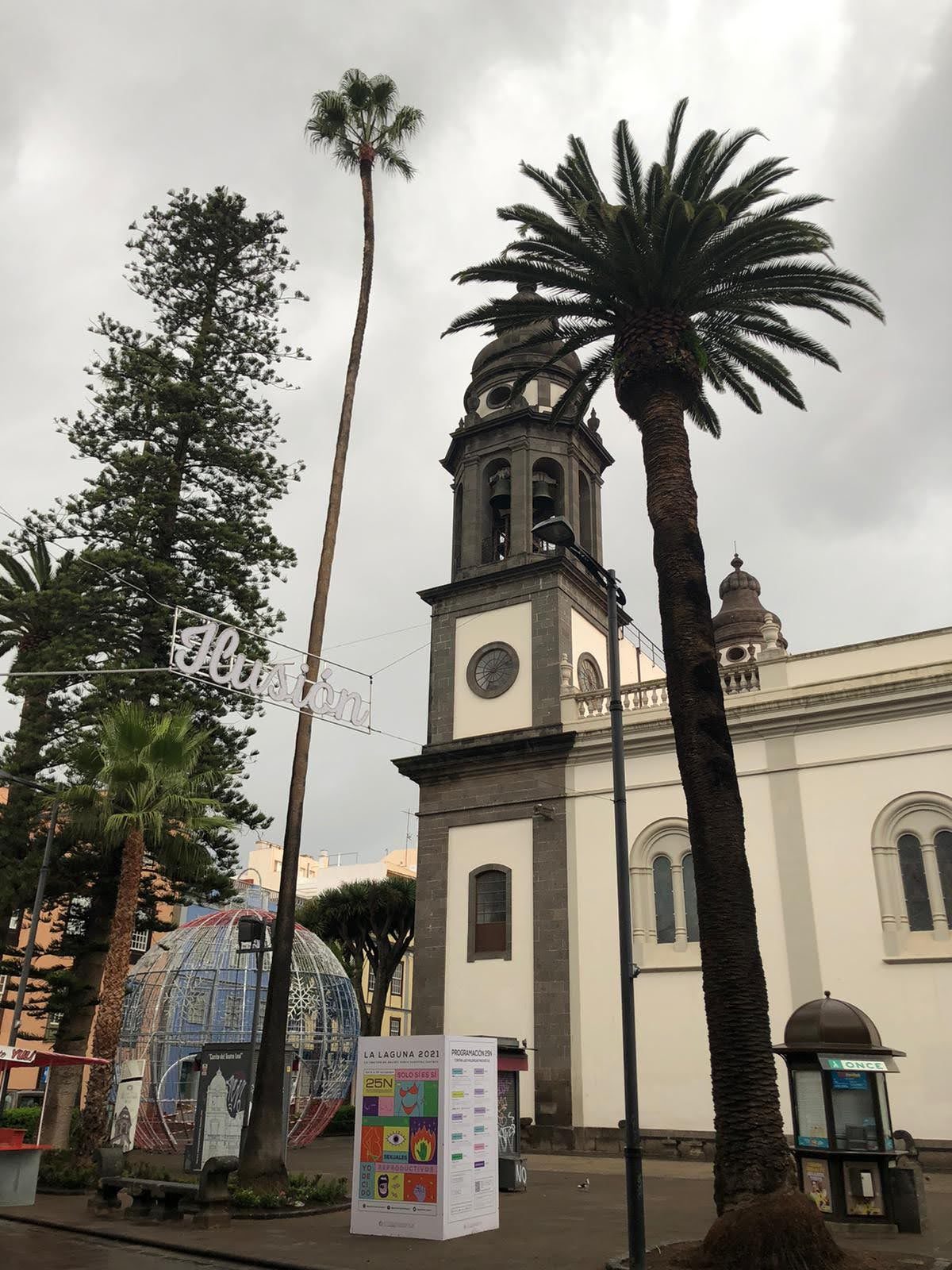 Torre de iglesia histórica rodeada de altas palmeras y adornos navideños bajo cielo nublado.