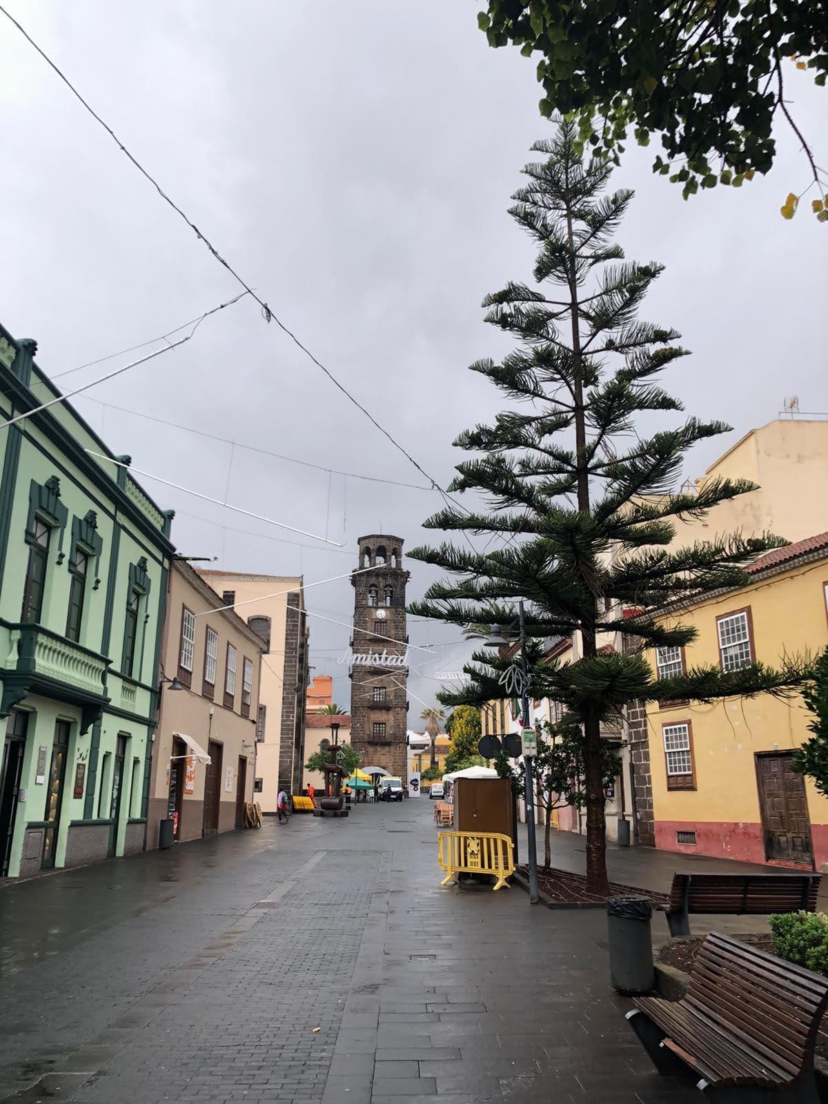 Calle peatonal húmeda entre edificios tradicionales con una torre campanario al fondo.