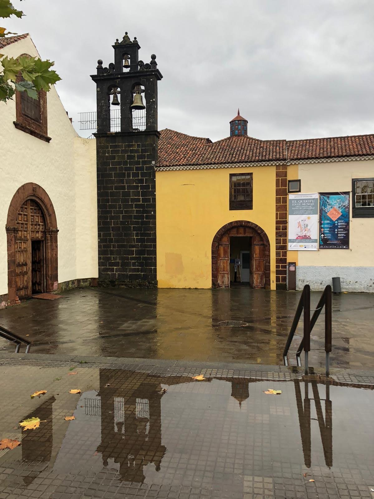 Campanario histórico reflejado en un charco de una plaza mojada tras la lluvia.