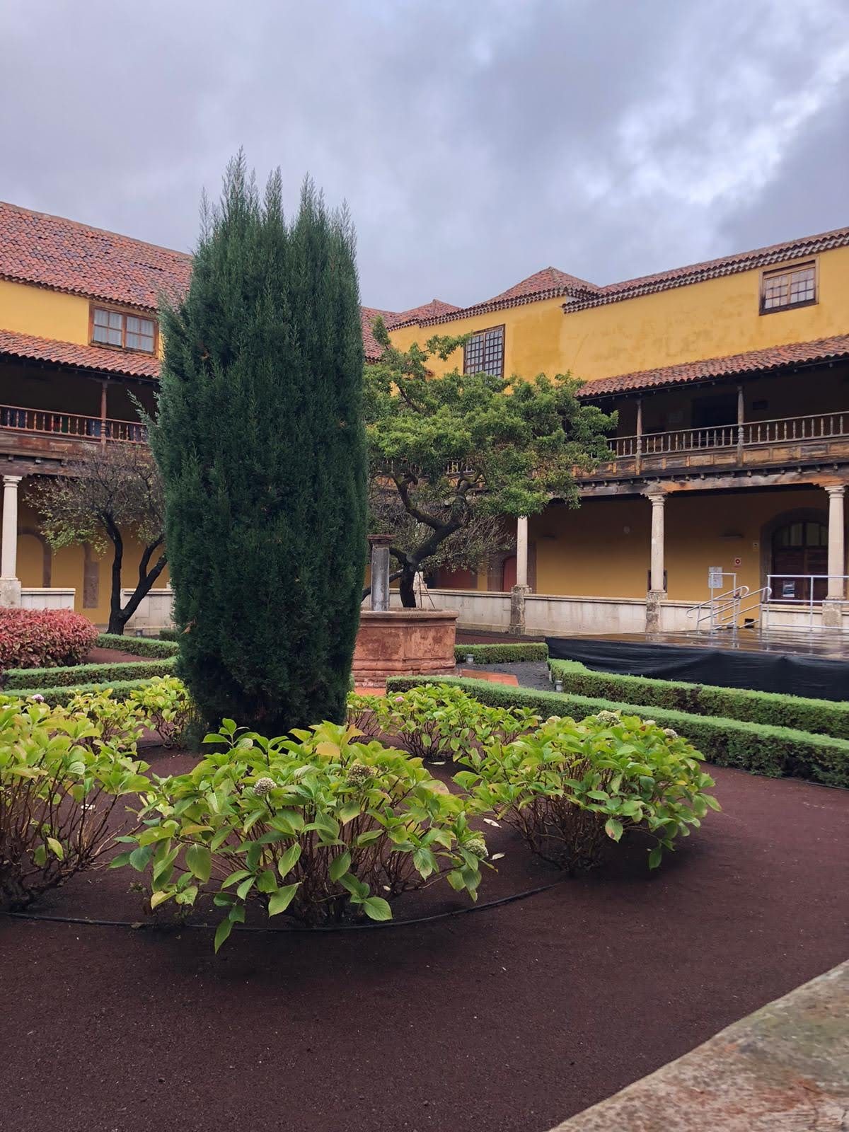 Patio interior tradicional canario con ciprés, jardines cuidados, fuente y balconadas de madera.