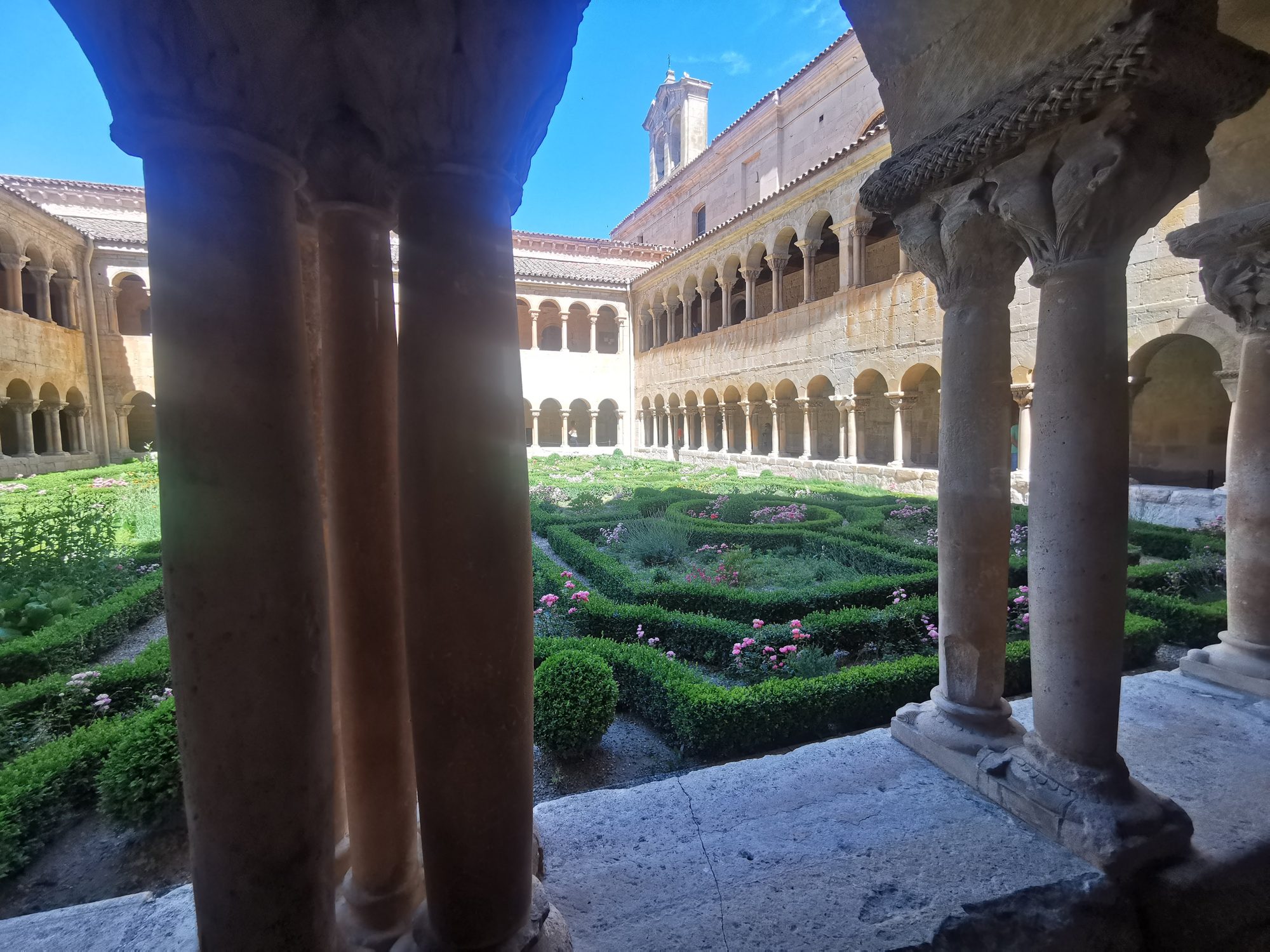 Claustro románico con columnas talladas y jardín interior con setos laberínticos y flores.