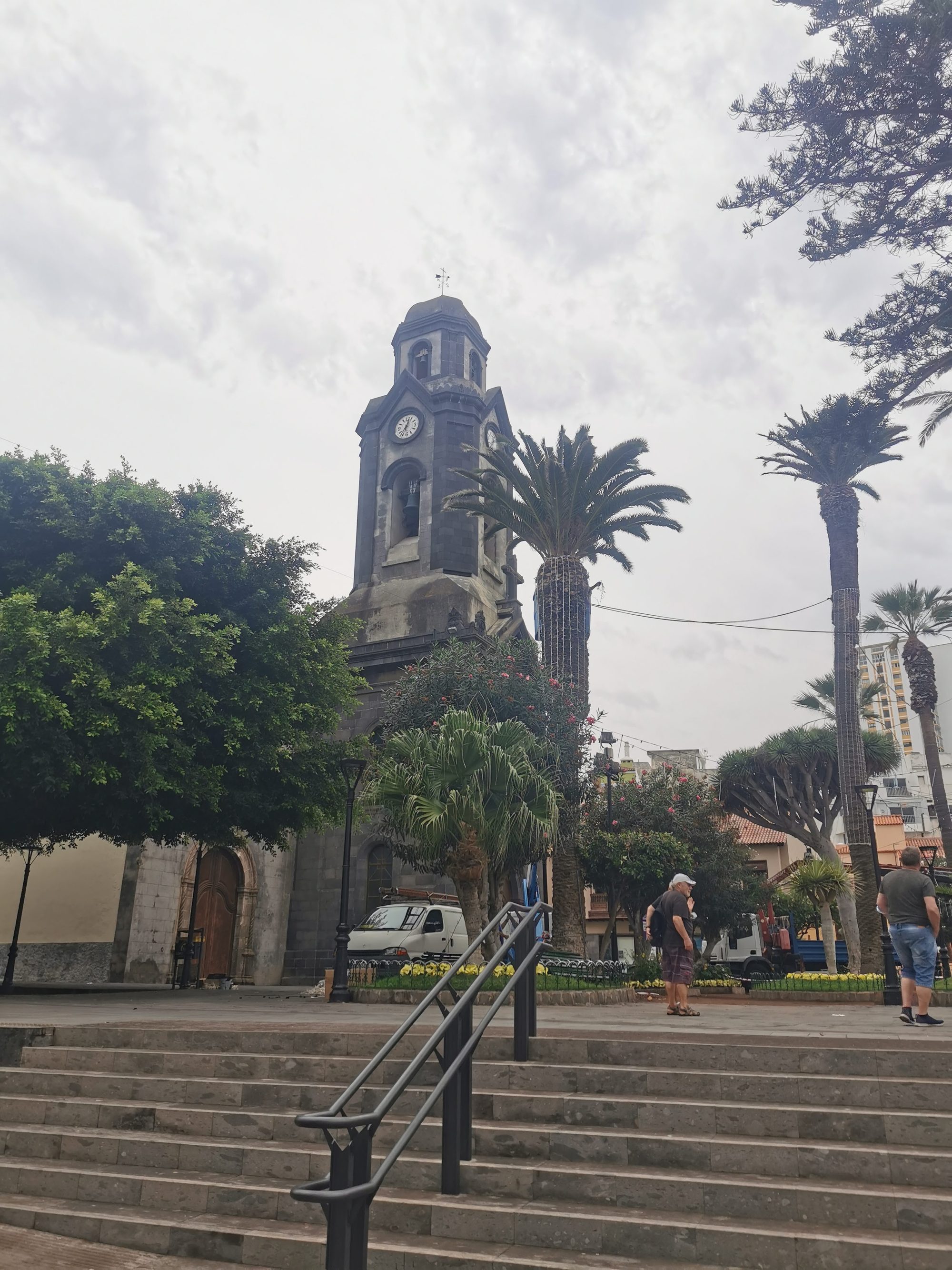 Torre campanario de piedra rodeada de palmeras en una plaza con escaleras.