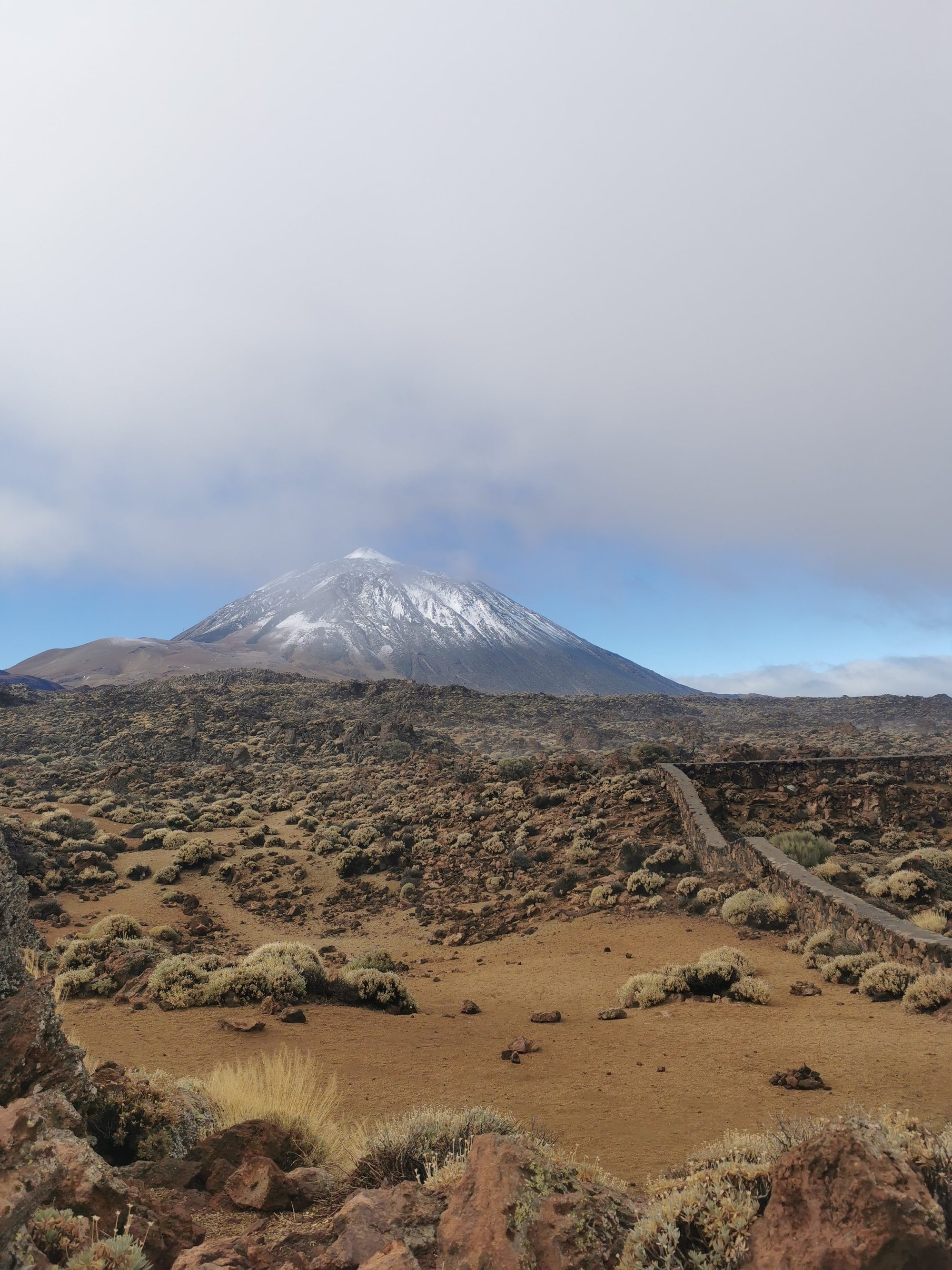 Vista del volcán Teide nevado sobre un paisaje volcánico árido y cielo nublado.